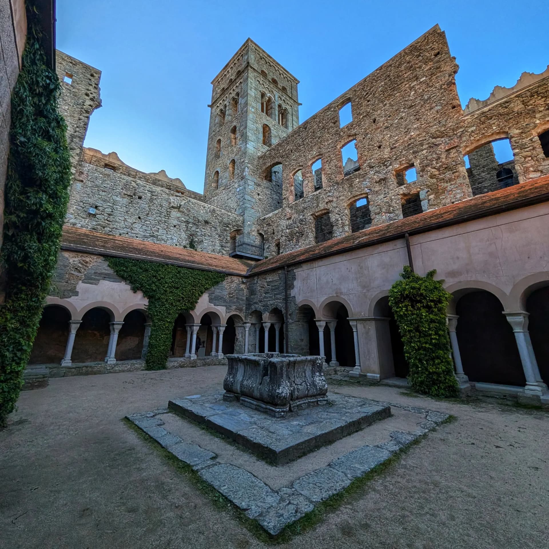 Pati interior del Monestir de Sant Pere de Rodes amb claustres romànics, torre i pou de pedra. Cel blau clar.