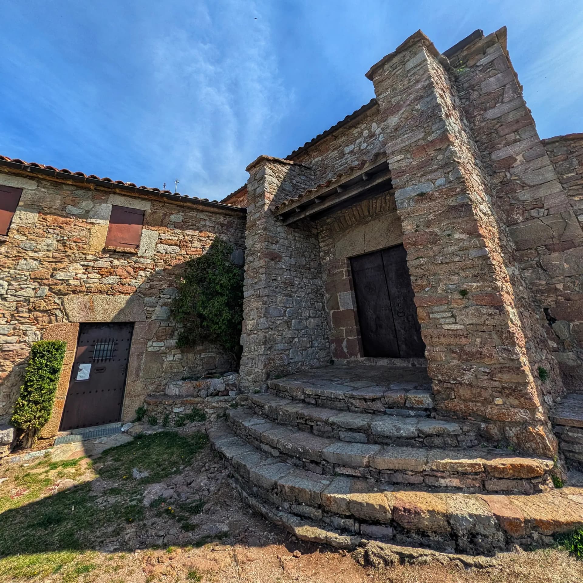 Antiga ermita de pedra amb murs envellits de tons rogencs i marrons, entrada d'arc, graons corbats i cel blau clar.