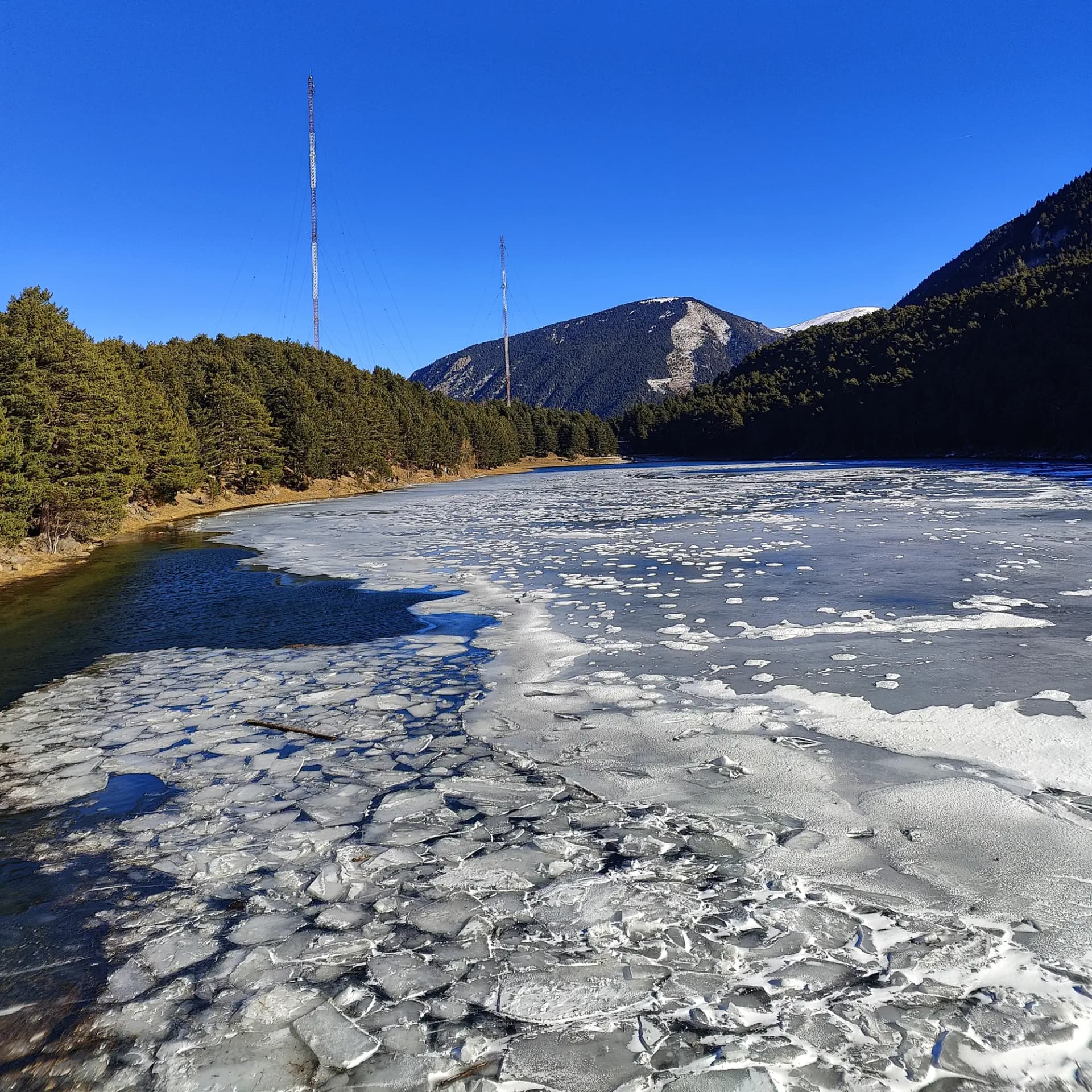 Partially frozen mountain lake with broken ice, pine forests, snowy peaks, and two communication towers.