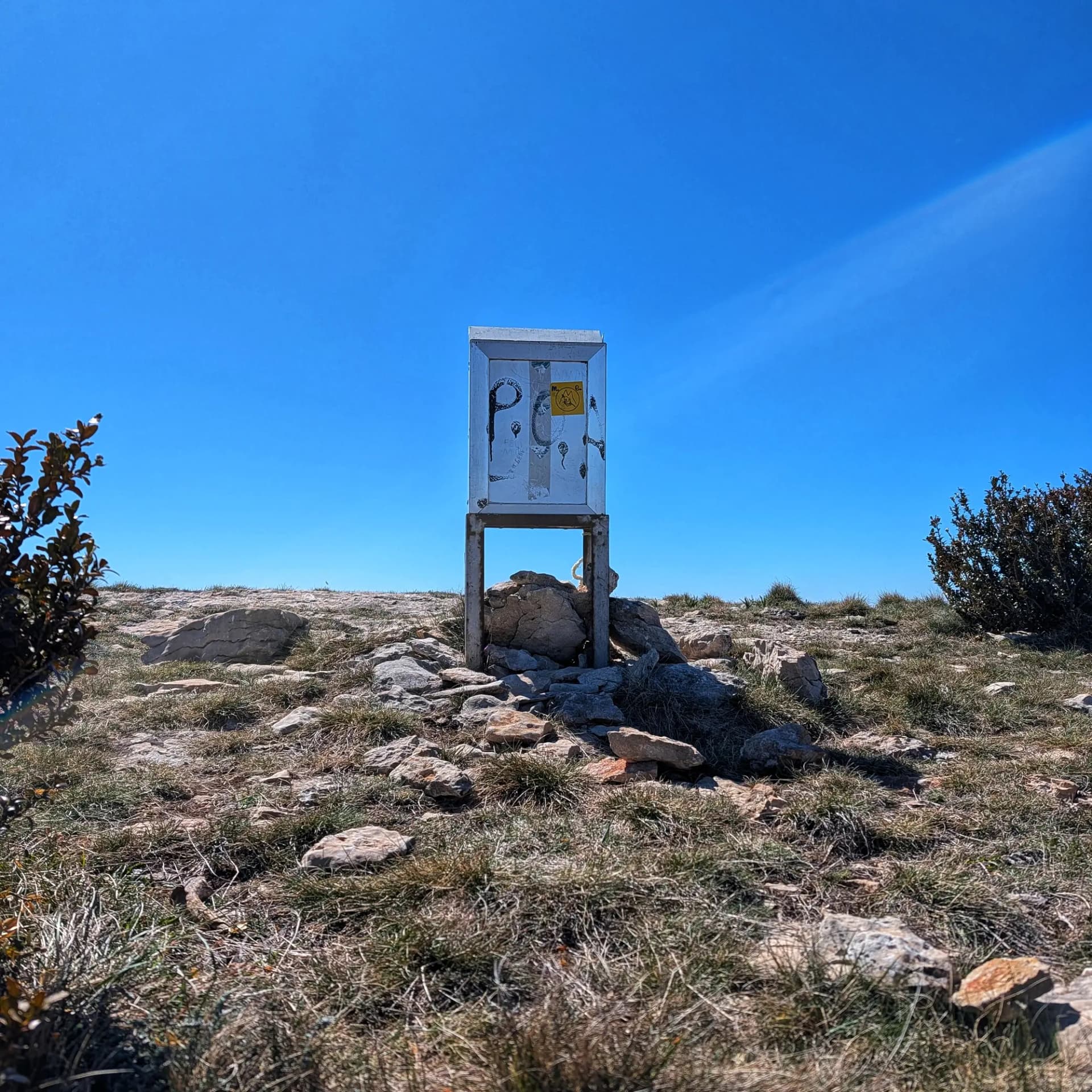 Caja metálica con grafitis en la cima del Tossal de Mirapallars, bajo un cielo azul claro.