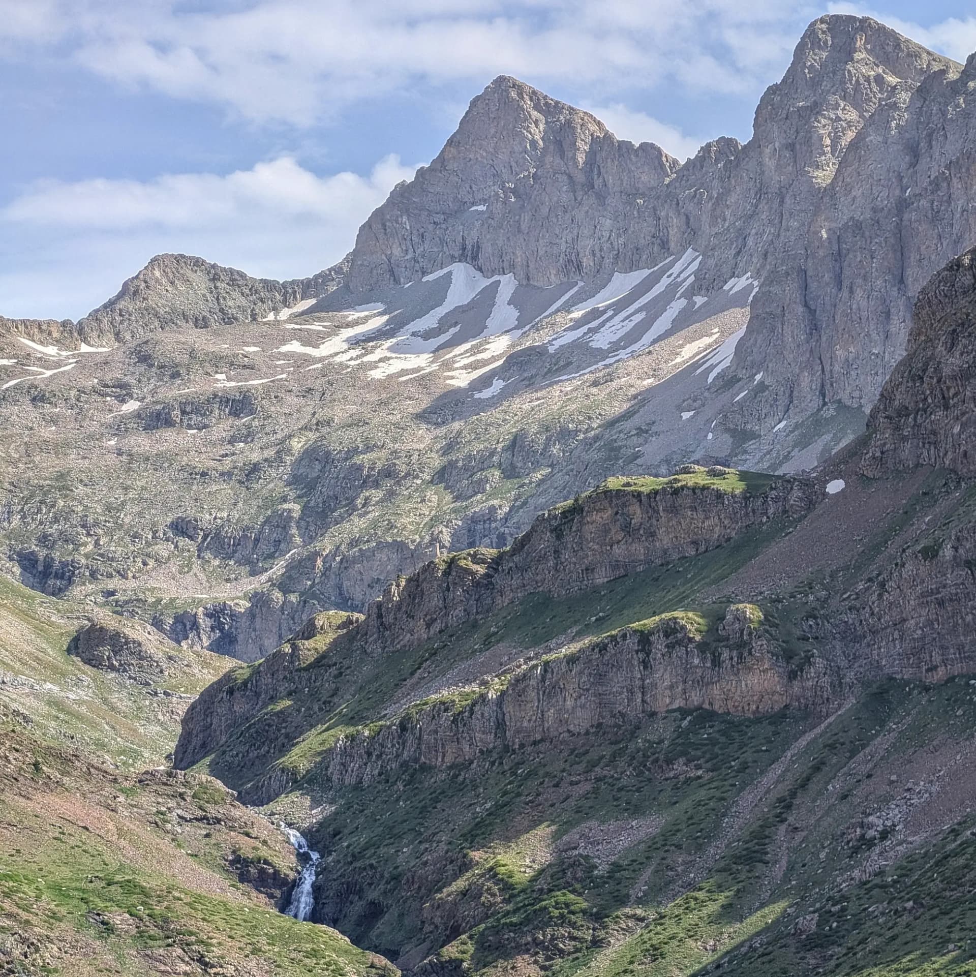 Paisatge alpí dramàtic amb cims rocosos escarpats, clapes de neu, i una cascada baixant per una vall verda sota cel blau.