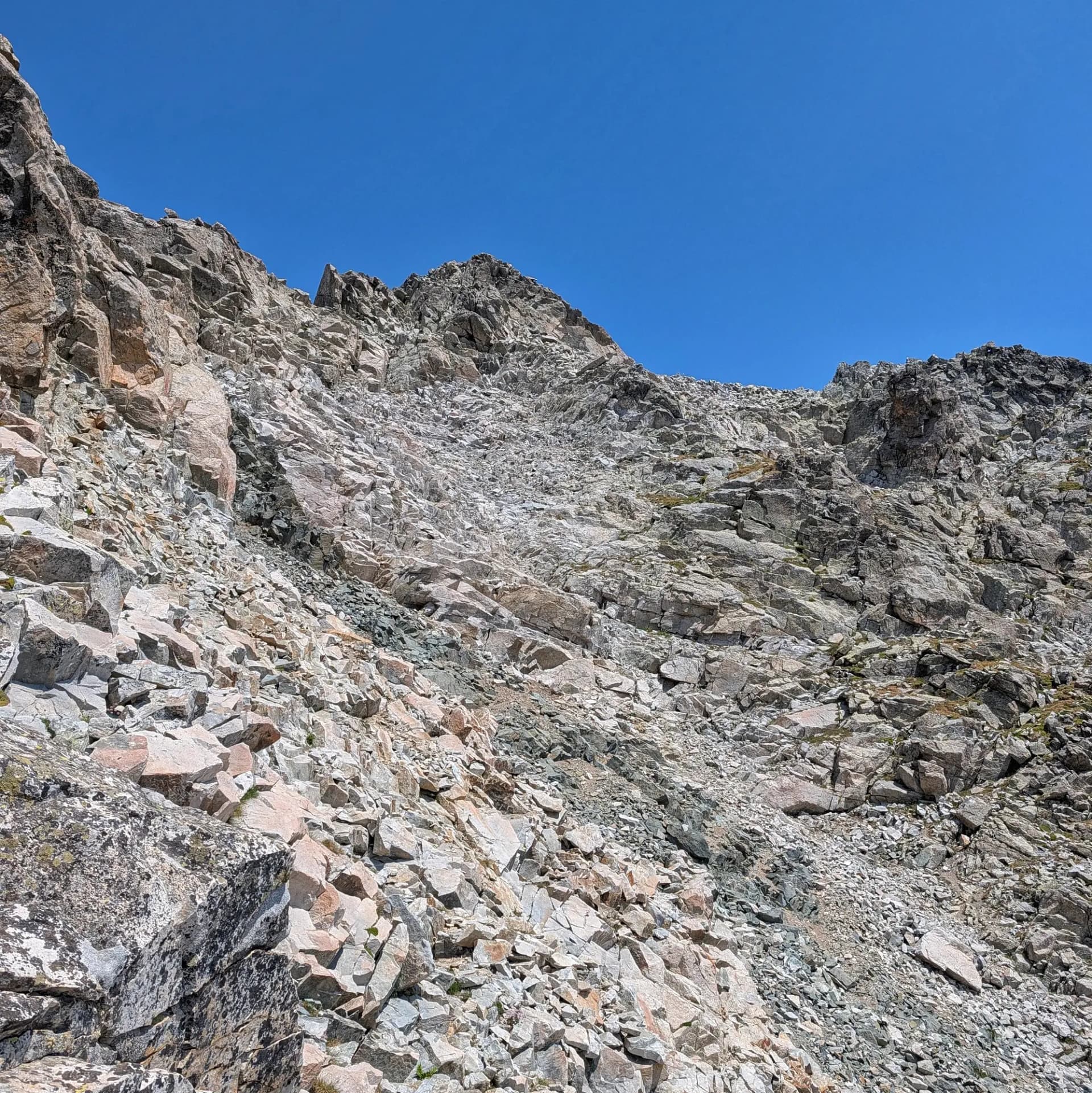 Faldilla rocosa i escarpada de muntanya amb roques grises i marronoses sota un cel blau clar, vista des de baix.