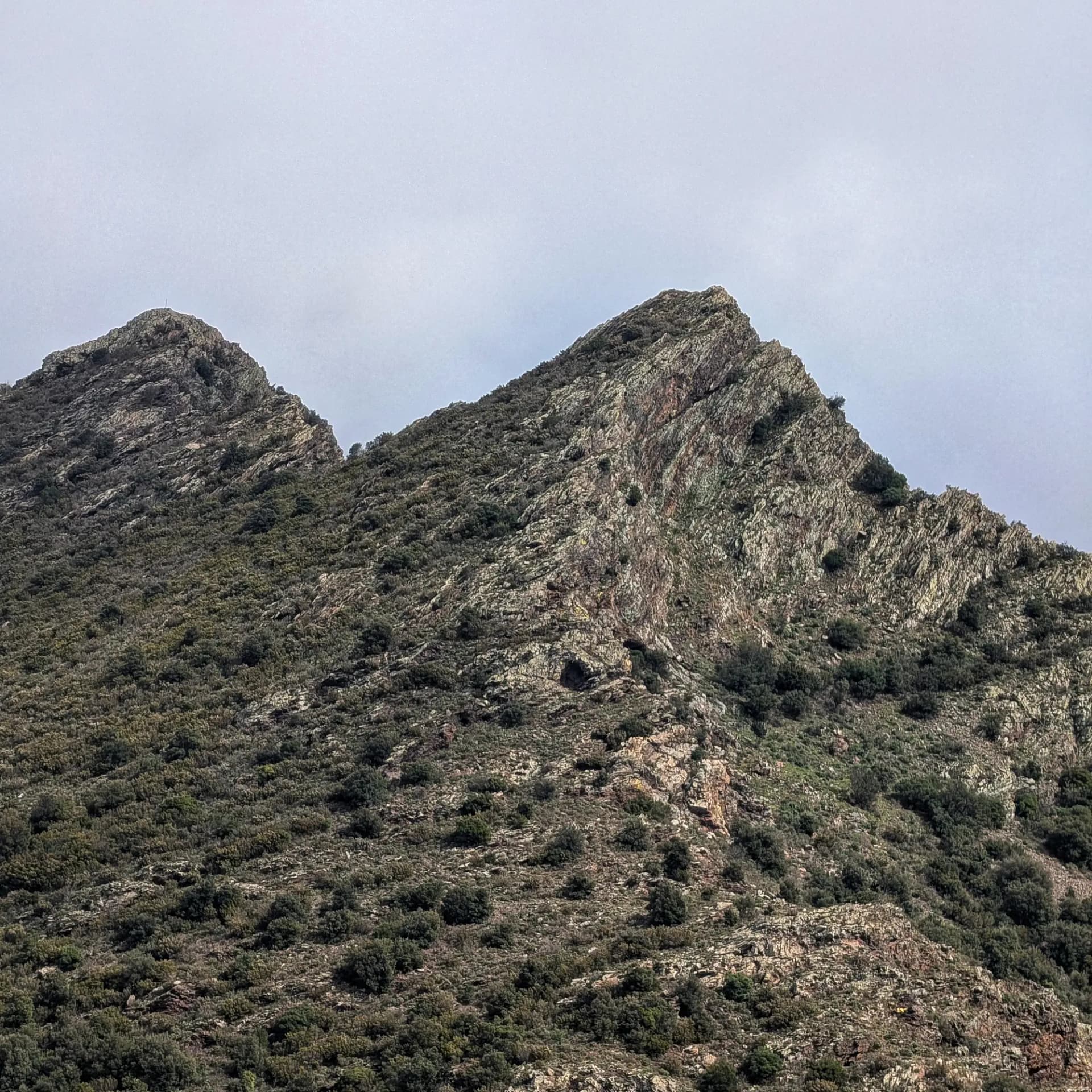 Crestas rocosas de las Orejas de la Mula con vegetación escasa y cruz en la cima, bajo cielo gris.