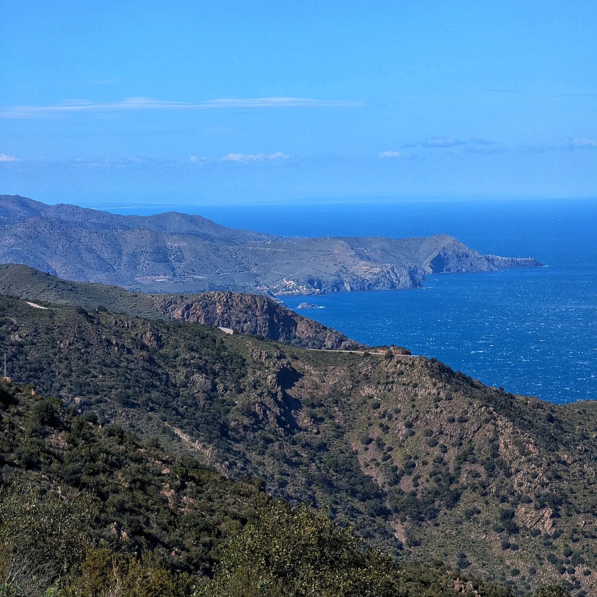 Panoramic coastal view with green hills, rocky cliffs, and a deep blue, choppy sea under a clear sky.