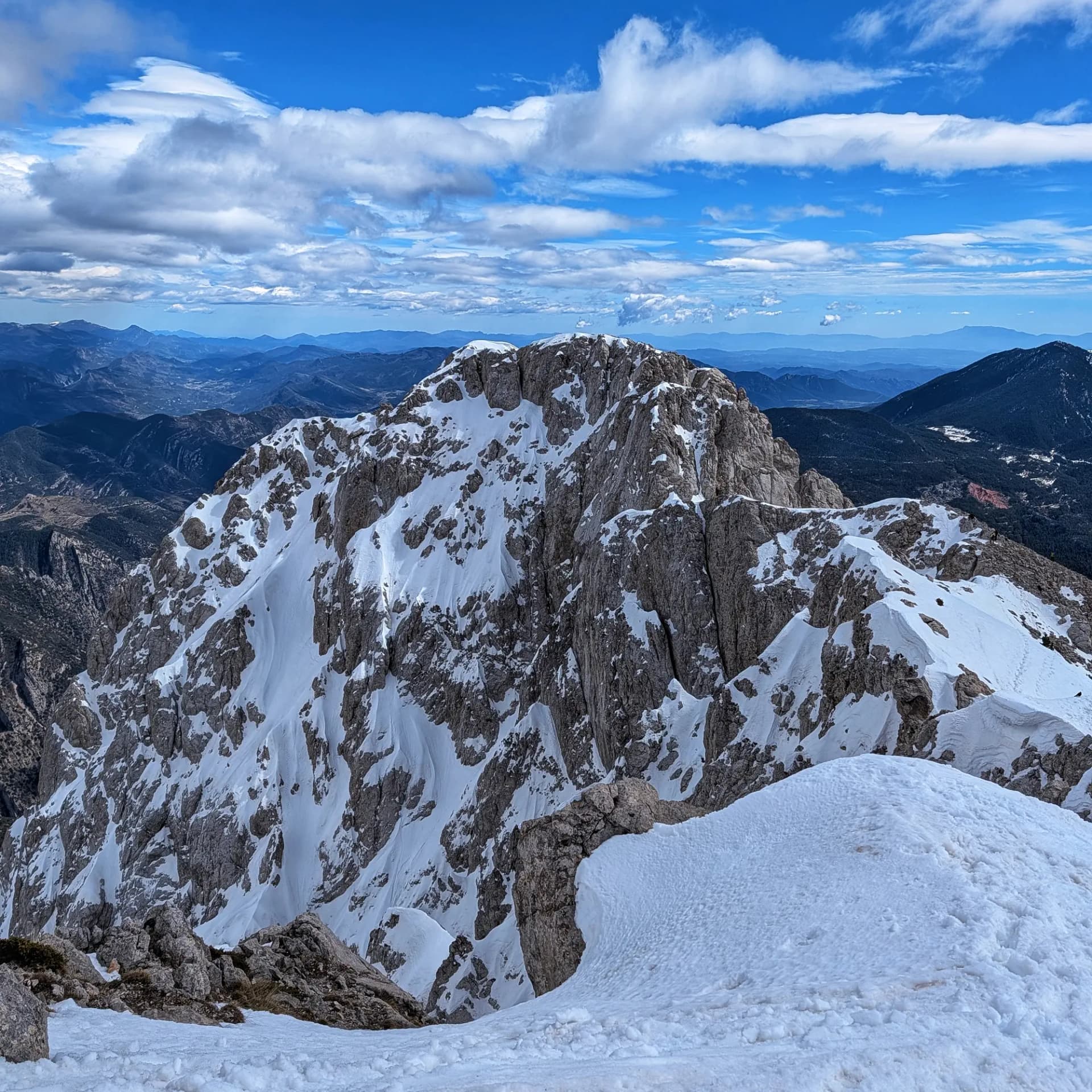 Paisatge muntanyós nevat. Un pic rocós cobert de neu en primer pla amb serralades llunyanes i cel blau amb núvols.