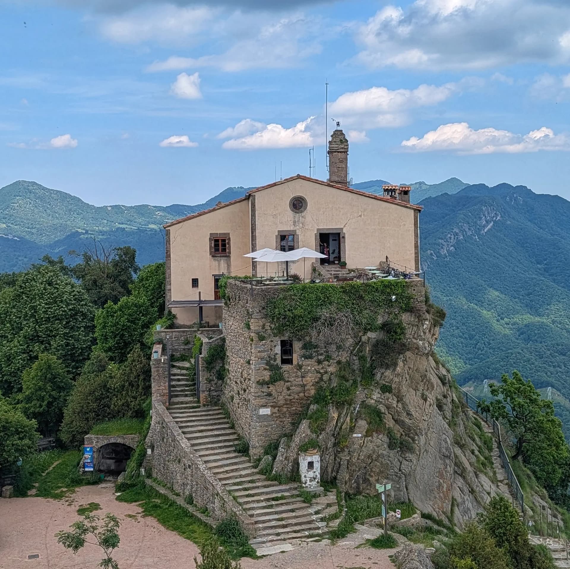 Santuari de Bellmunt building on a rocky cliff with stone stairs, green mountains under a blue cloudy sky.