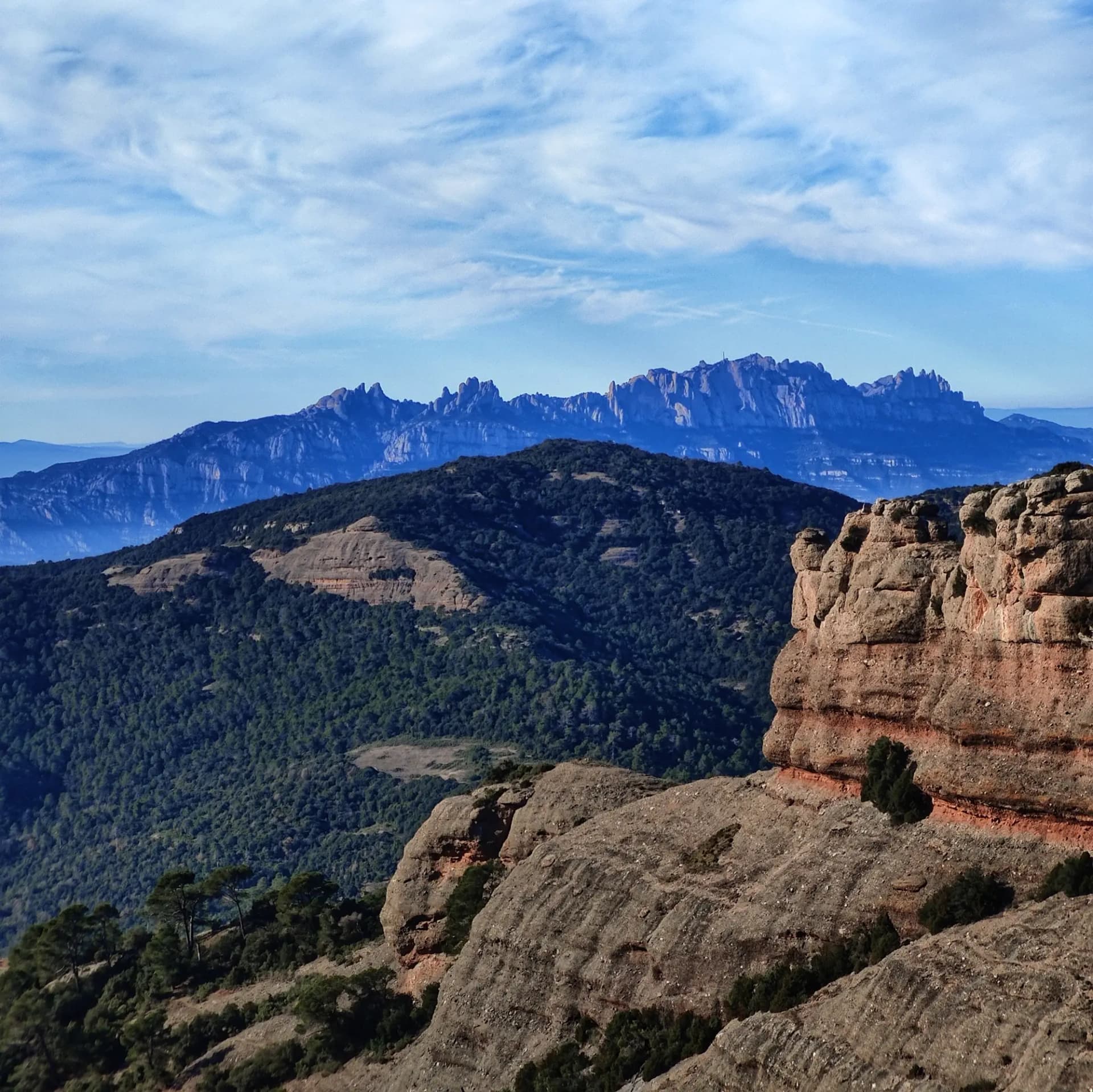 View from Sant Llorenç del Munt showing rugged brown rock formations, a green forest, and the distant jagged peaks of Montserrat under a blue sky.