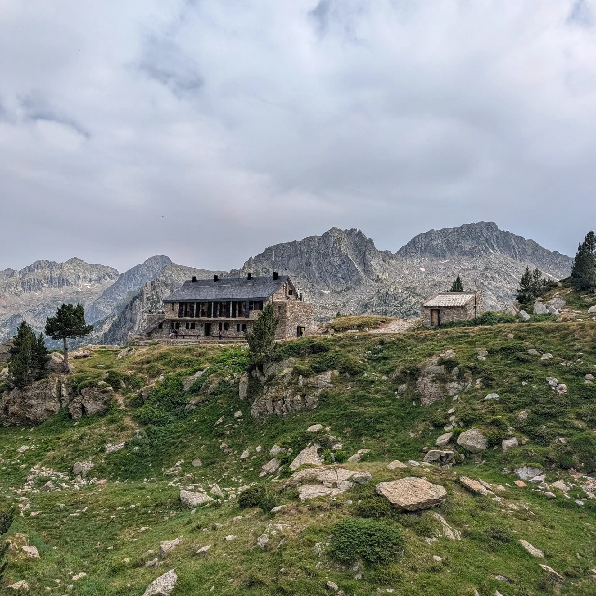 Refugi de muntanya de pedra i petita cabana en un vessant rocós i verd, amb pins i pics escarpats sota un cel ennuvolat.