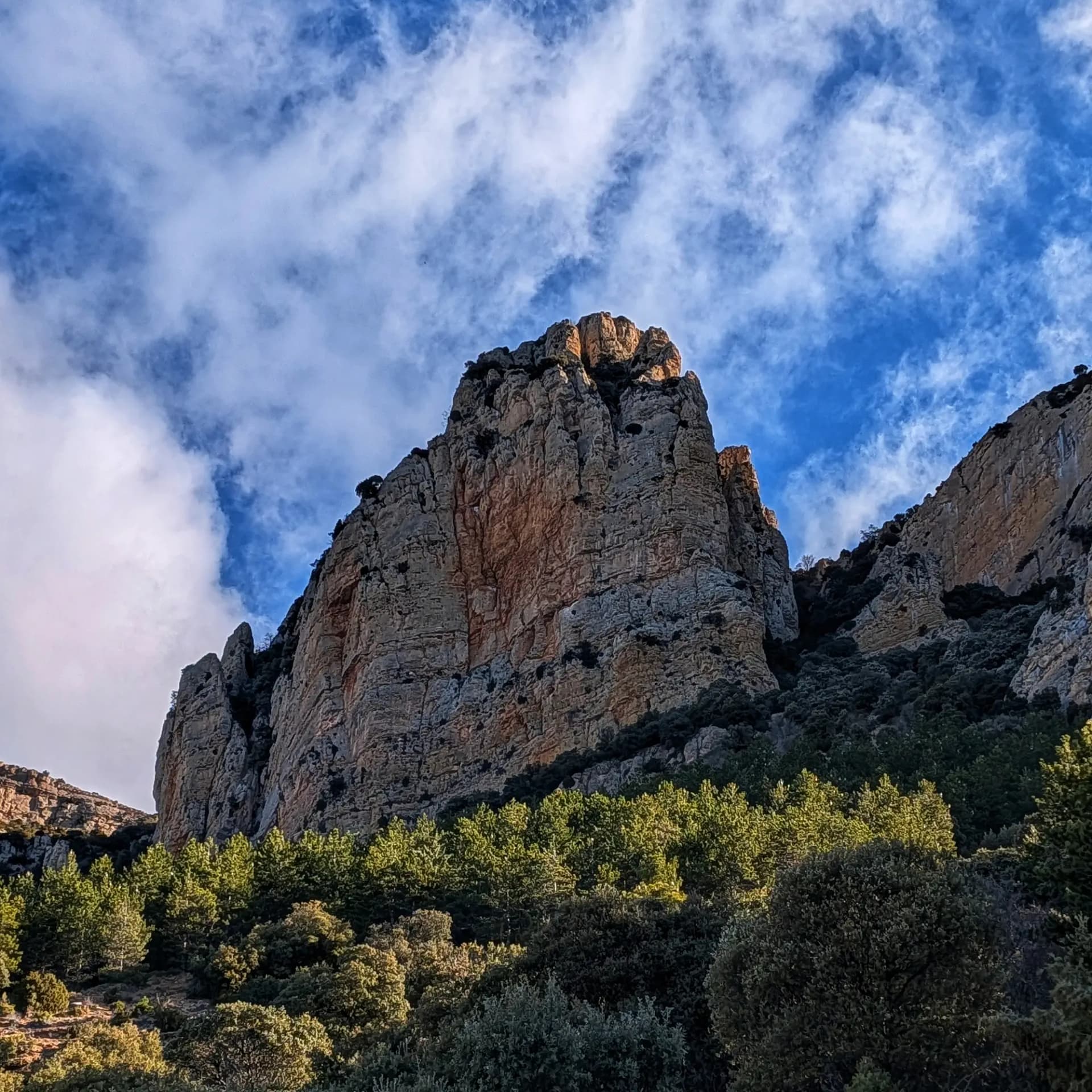 Imponente formación rocosa de Montsec de Rúbies iluminada por el sol, sobre un denso bosque verde y cielo azul con nubes.