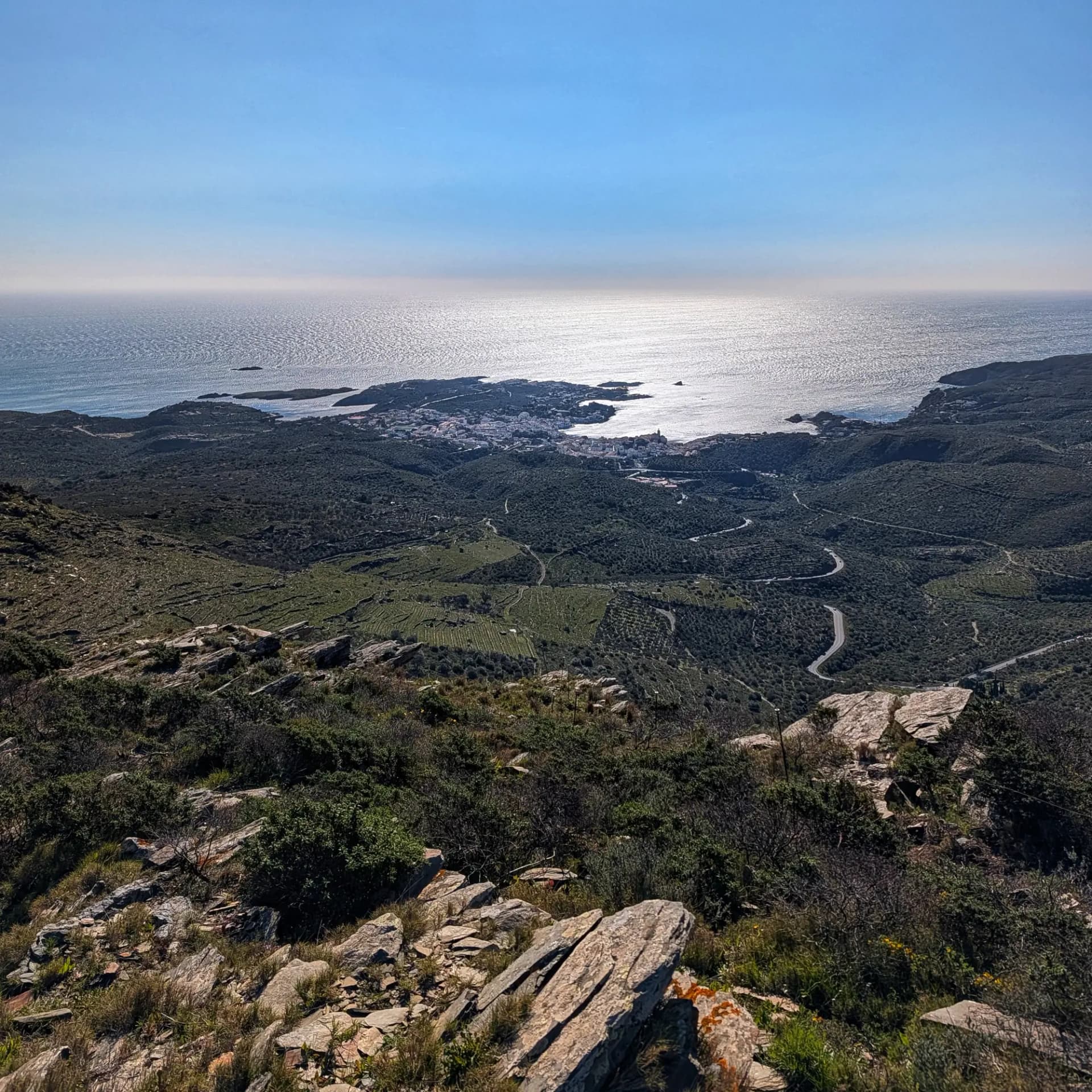 Panoramic view of Cadaqués town and the Mediterranean Sea from a rocky, vegetated hilltop on a sunny day.