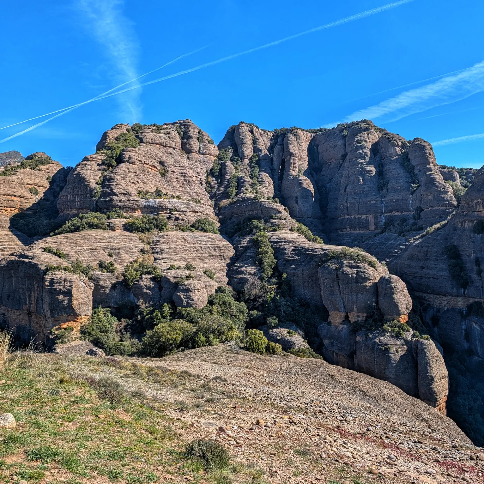 Formacions rocoses estratificades de color marró i verd sota un cel blau clar amb esteles de condensació.