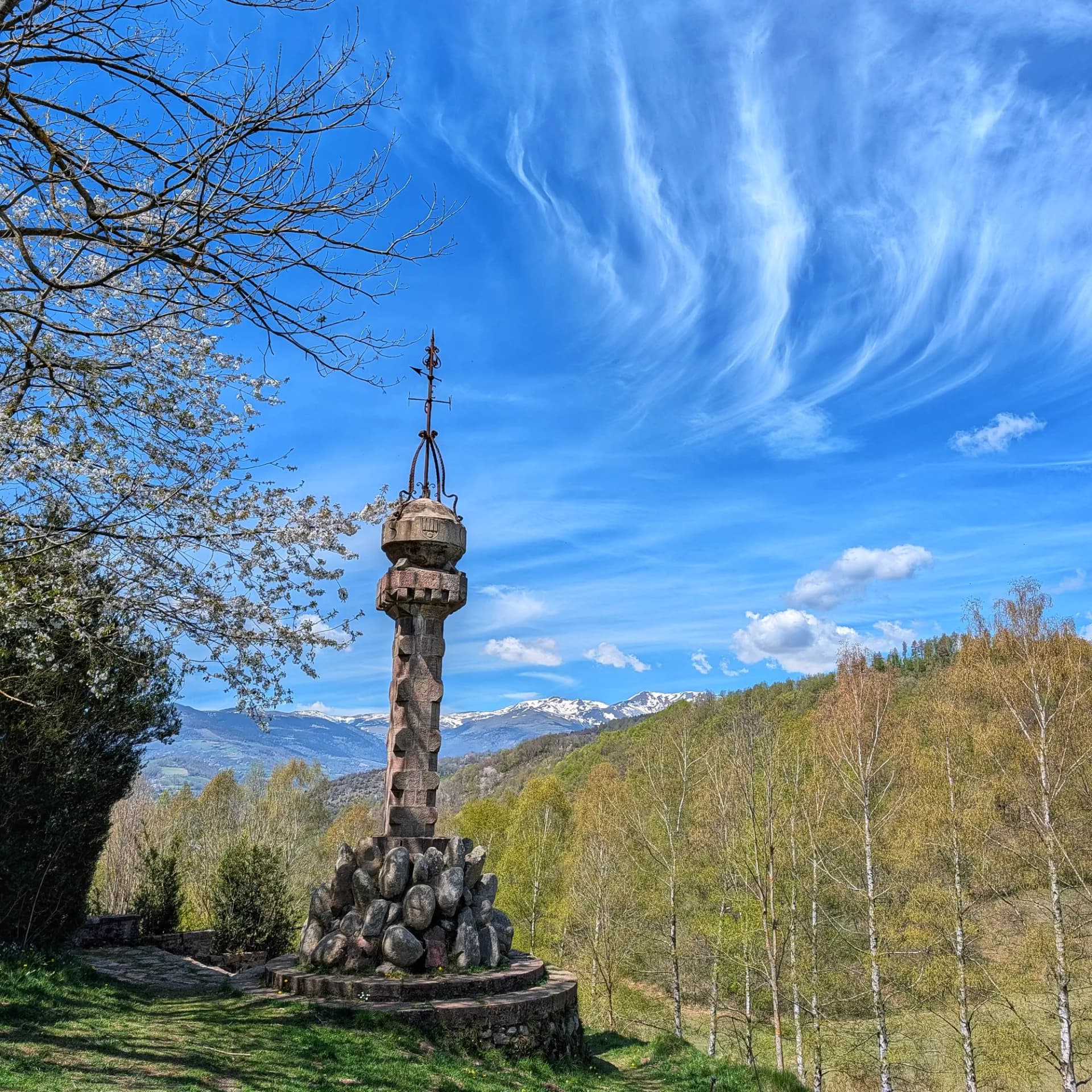 Monument de pedra amb pinacle metàl·lic sota cel blau, entre arbres de primavera i muntanyes nevades.
