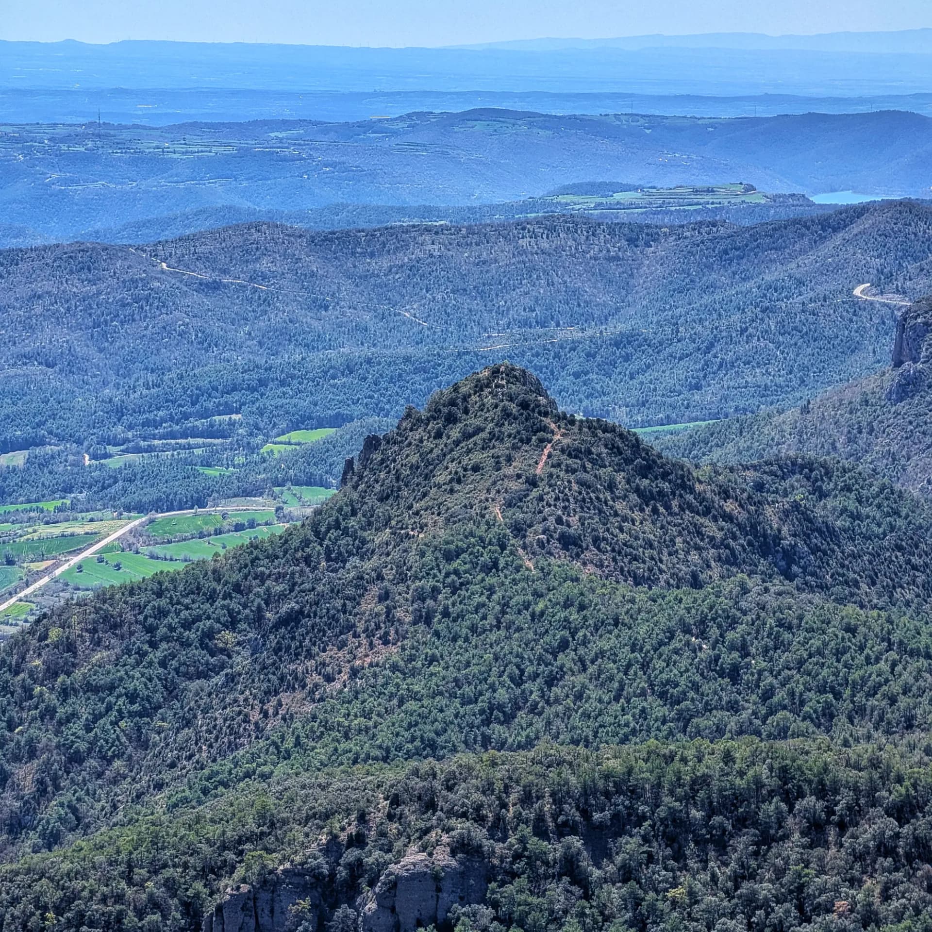 Vista aèria del Roc de Cogul, pic boscós envoltat de valls, turons distants i un embassament blau.
