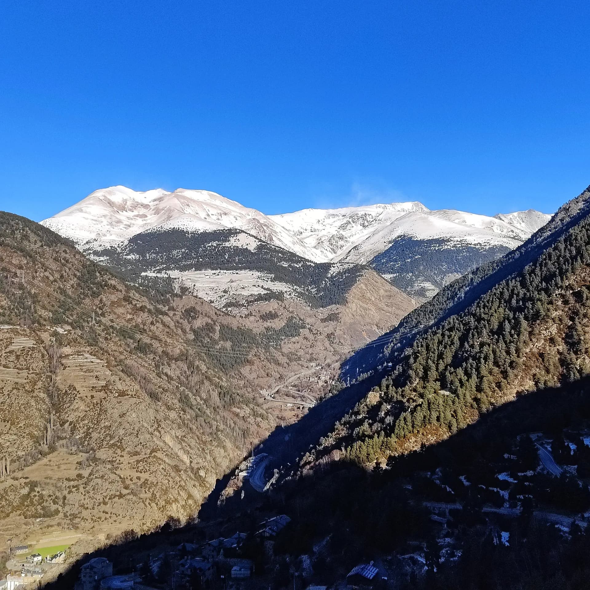 Snow-capped mountains and forested valleys under a clear, deep blue sky. Casamanya peak prominent in the distance.