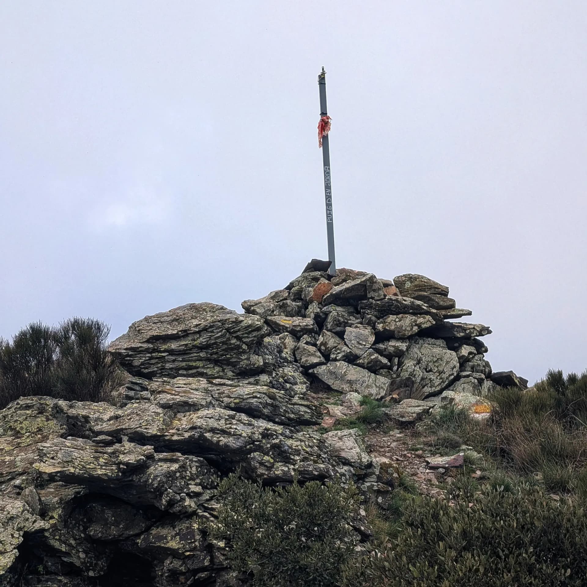 Cim rocós amb pal marcador metàl·lic amb la inscripció "PUIG D'EN JORDA" sota un cel ennuvolat.