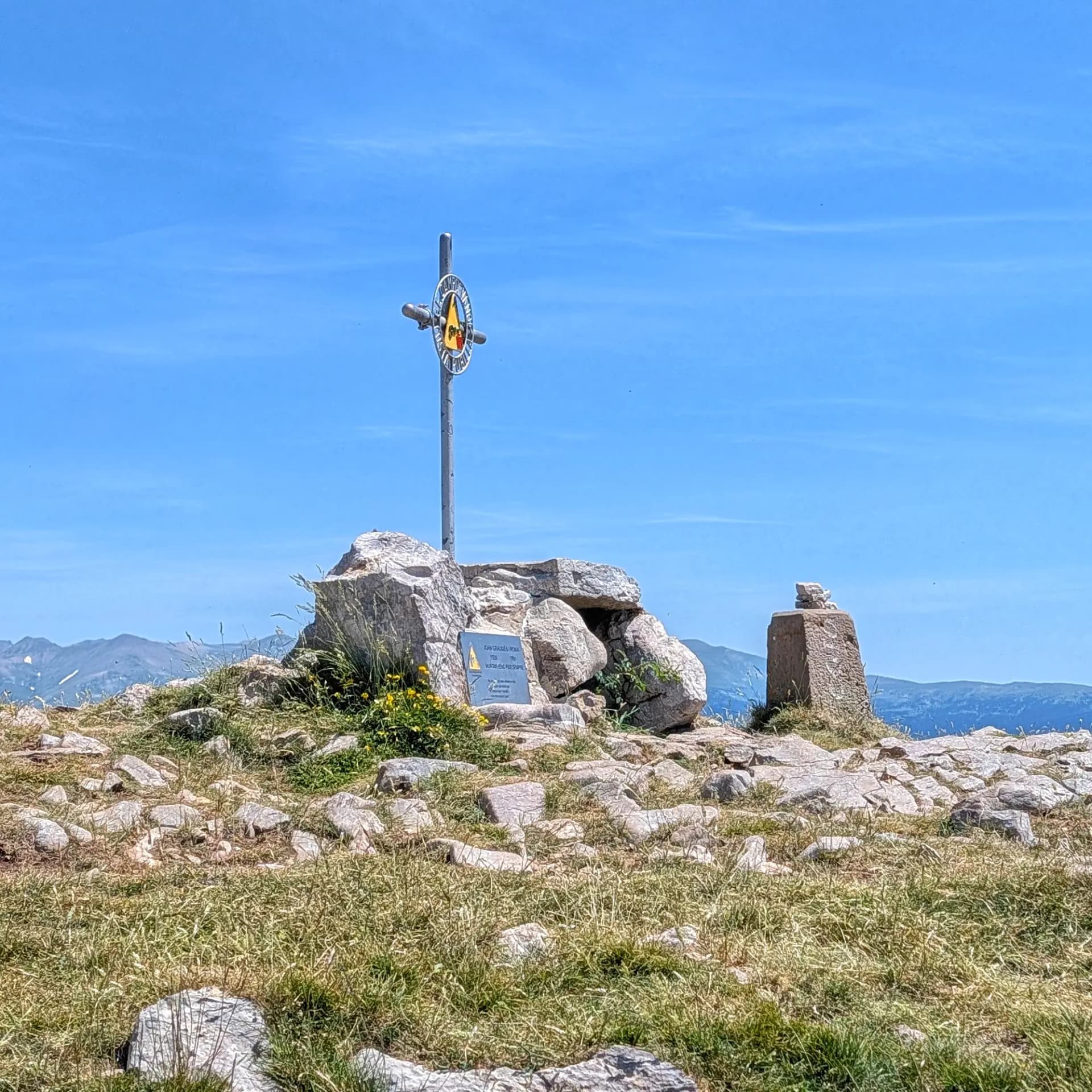 A clear view from the summit of Costa Pubilla (or Pla de Pujals), marked by a cross and stone pillar. This peak is often reached from the Santuari de Montgrony. Mountain summit with a metal cross marker, stone pillar, and plaque on a rocky, grassy peak under a clear blue sky.