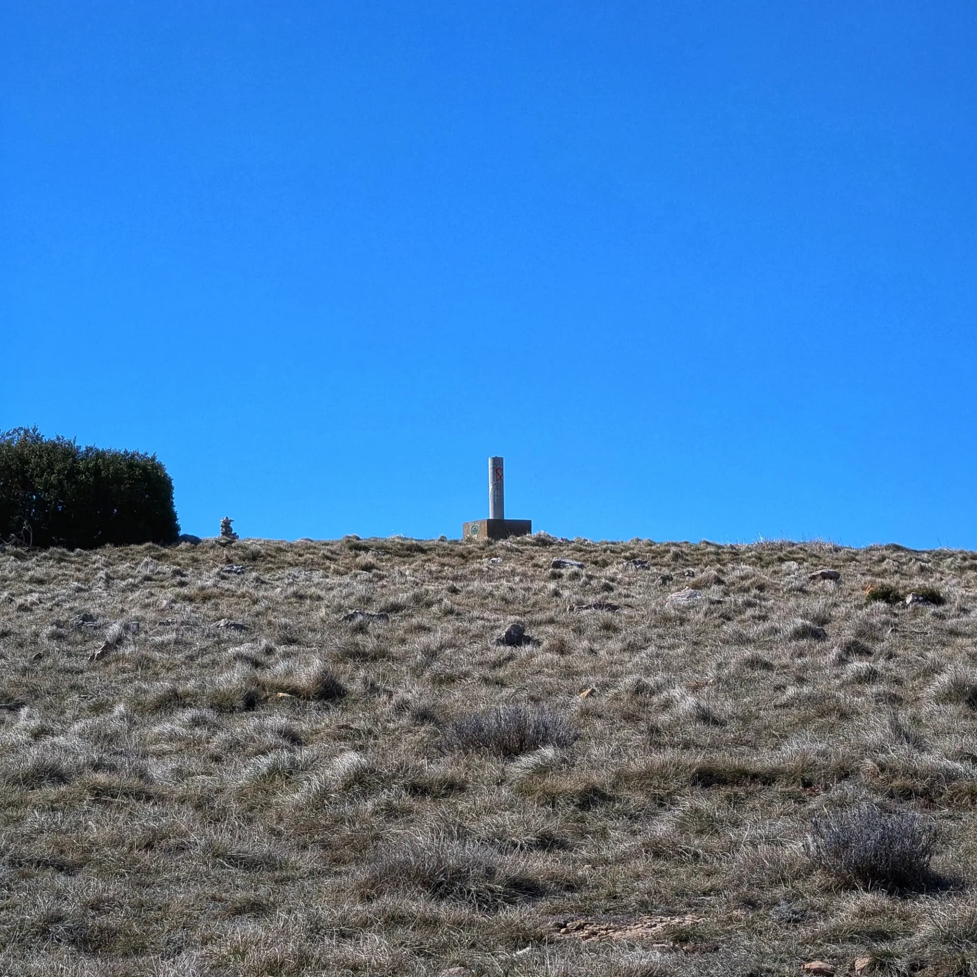 Hito geodésico en la cima del Tossal de les Torretes con hierba seca y cielo azul claro. Un mojón y arbusto a la izquierda.