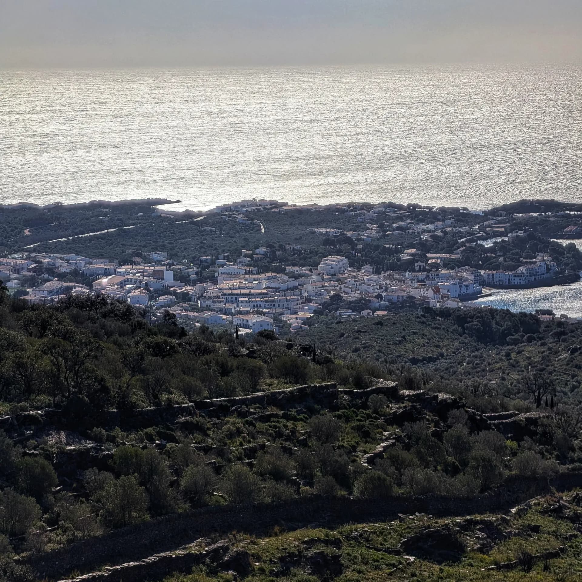 High-angle view of a white coastal town nestled between green hills and a sparkling sea under bright sky.