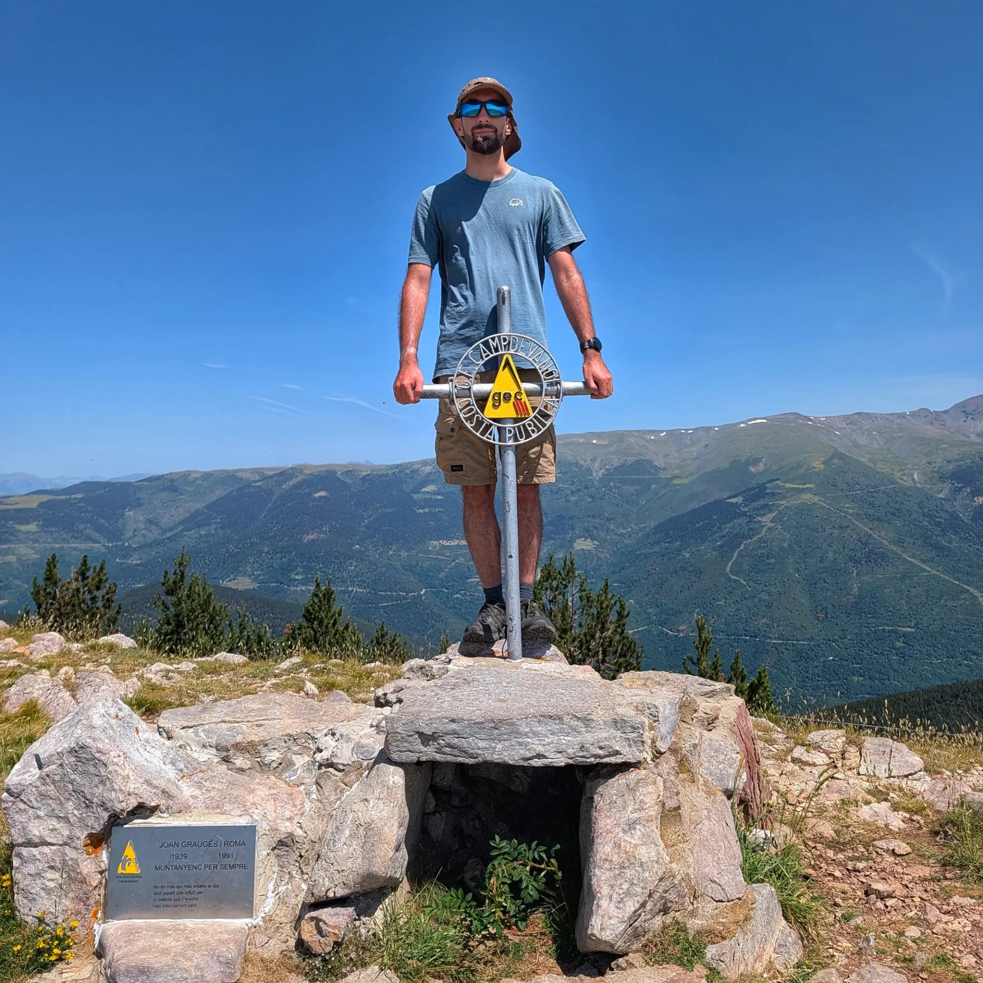 Costa Pubilla Man on a rocky mountain peak, holding a metal summit marker, with a vast green mountain range under a clear blue sky.