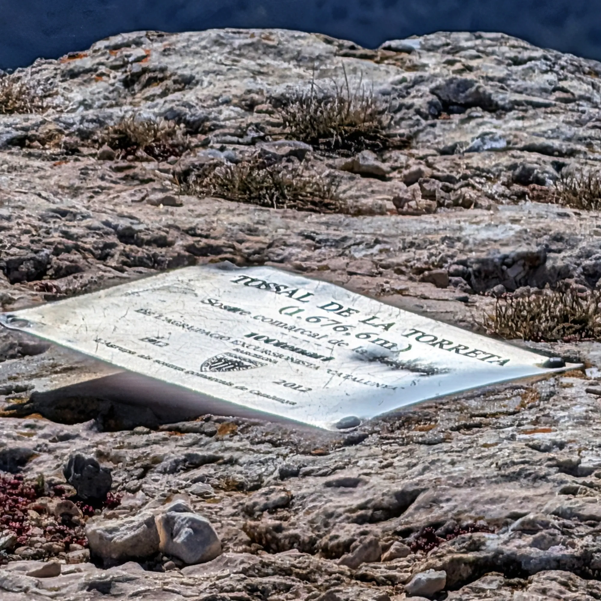 Placa metálica en la cumbre del Tossal de la Torreta (1676.6m) sobre terreno rocoso y árido.