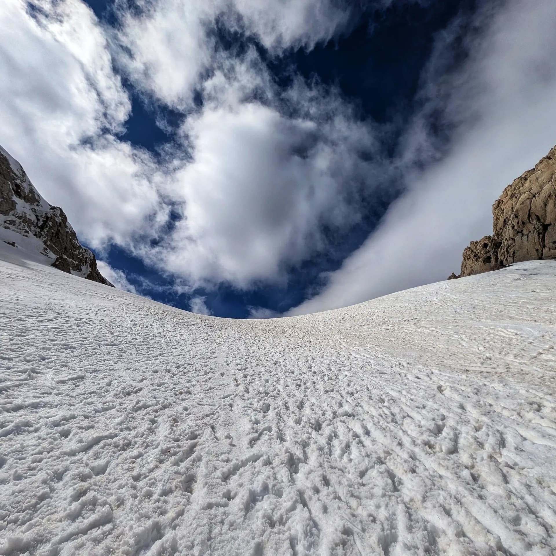 Pendís nevat de muntanya sota un cel blau intens amb núvols blancs i cims rocosos als costats.