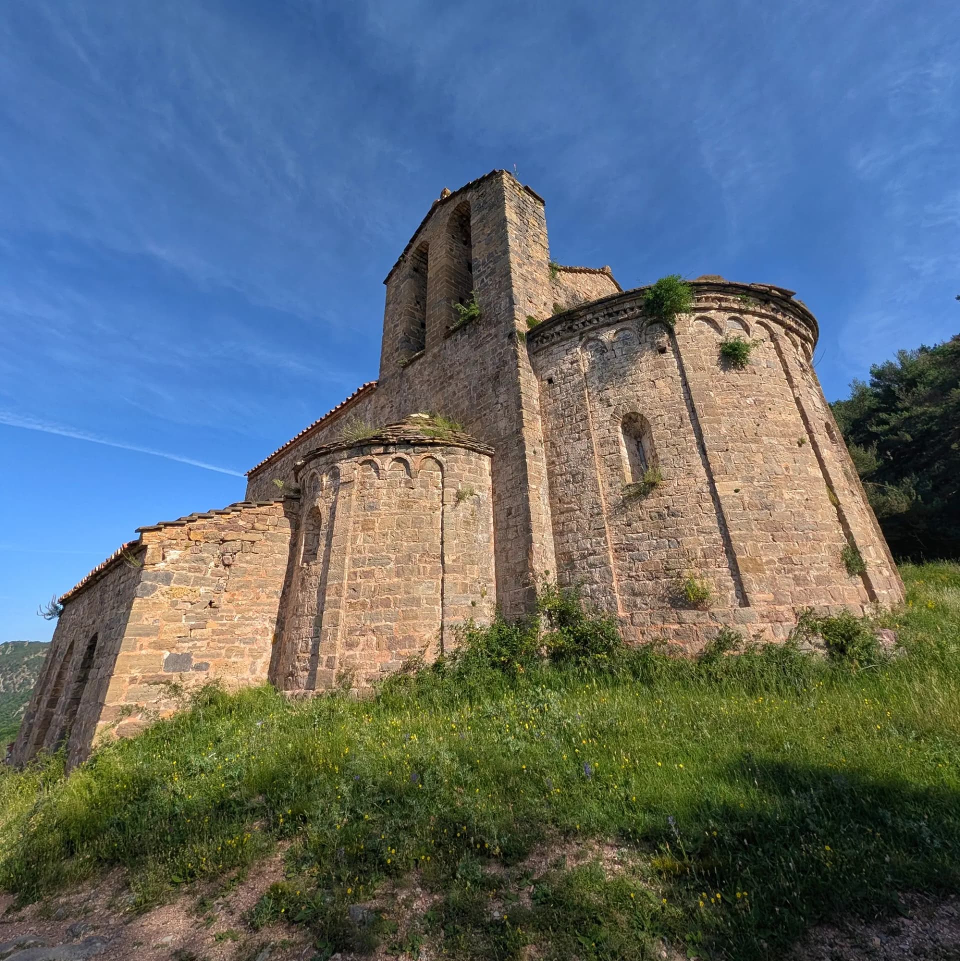 The historic Romanesque Sanctuary of Montgrony in the Catalan Pre-Pyrenees, a beautiful starting point for hikes to Costa Pubilla or Pla de Pujals. Historic Romanesque stone church with a bell tower and rounded apse on a green, grassy hill under a bright blue sky.