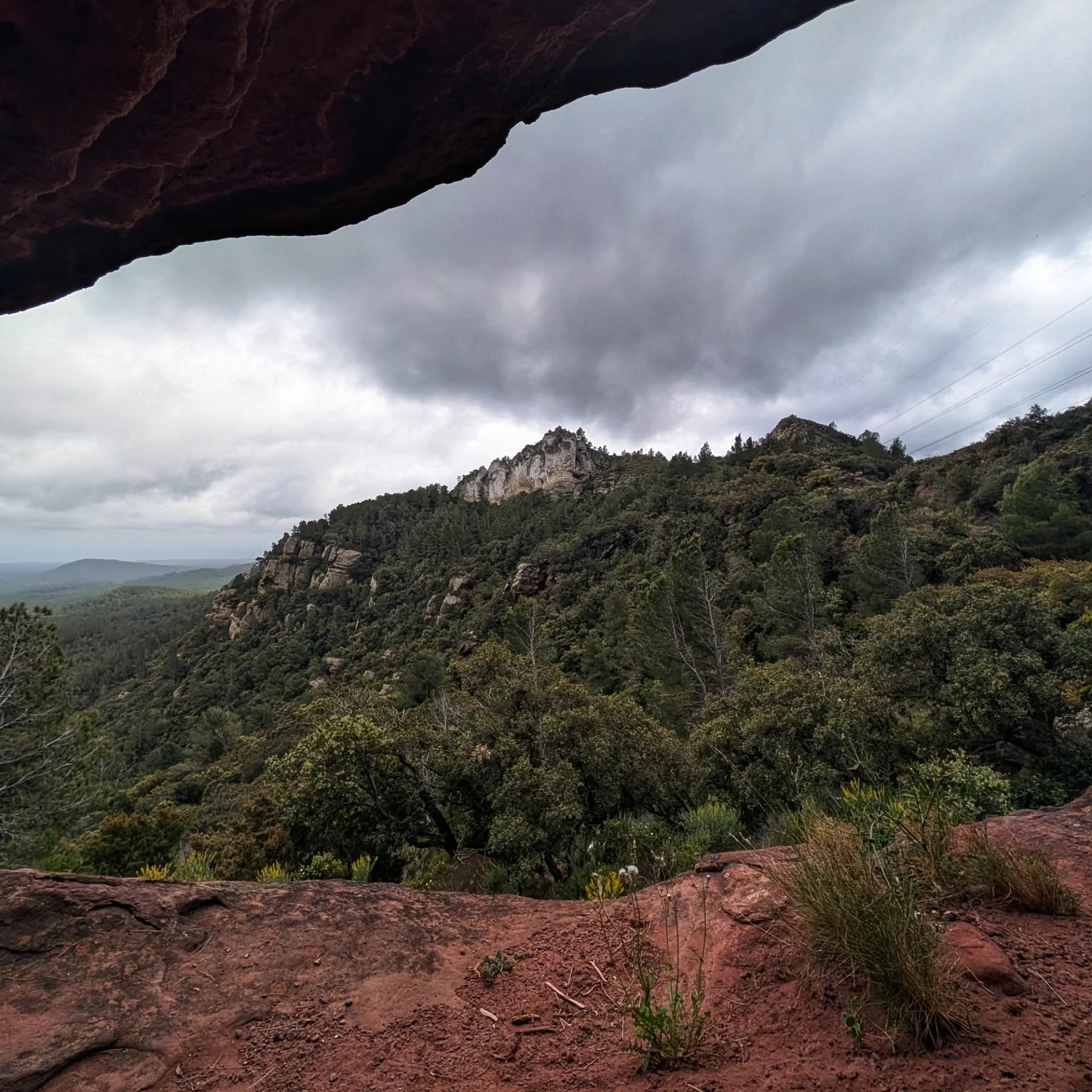 Vista panoràmica d'un paisatge muntanyós boscós sota cel ennuvolat, emmarcada per l'obertura vermellosa d'una cova de Buntsandstein.