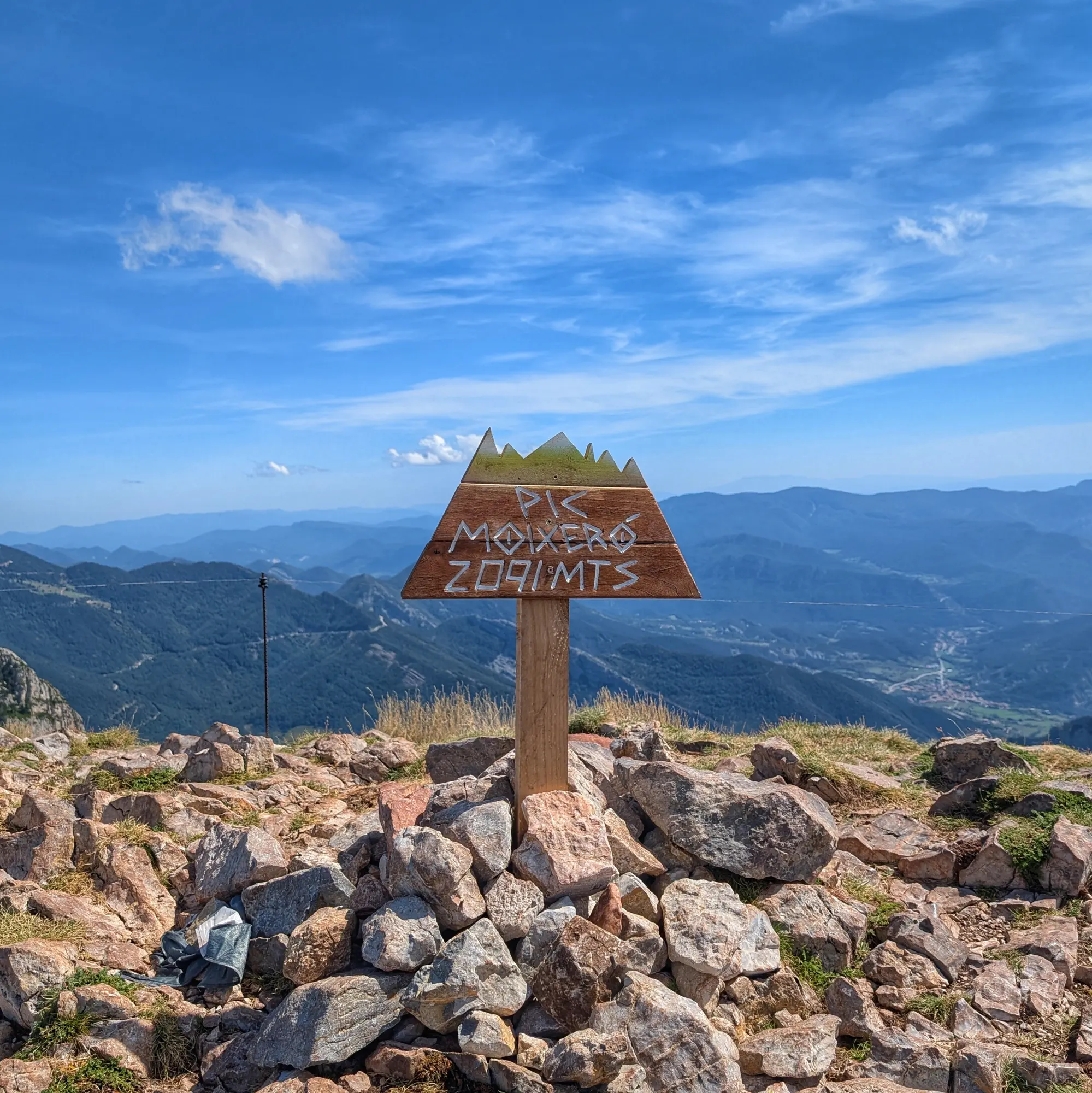 Triangular wooden signpost at the Pic del Moixeró summit with the inscription "Pic Moixeró 2,090 mts" under a blue sky.