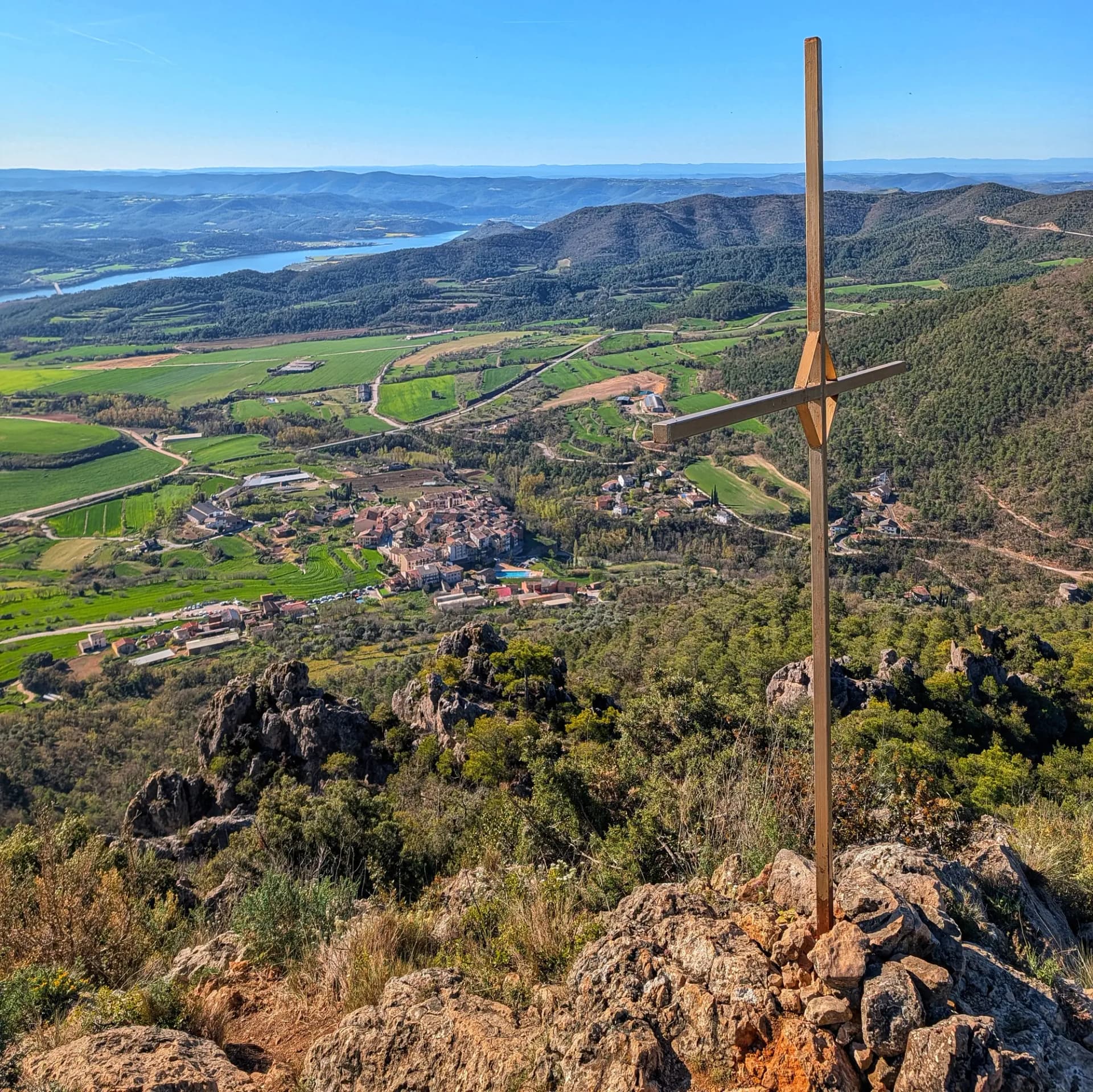 Vista panoràmica des del Roc de Cogul amb el poble de Peramola, camps verds, el Pantà de Rialb i una creu.