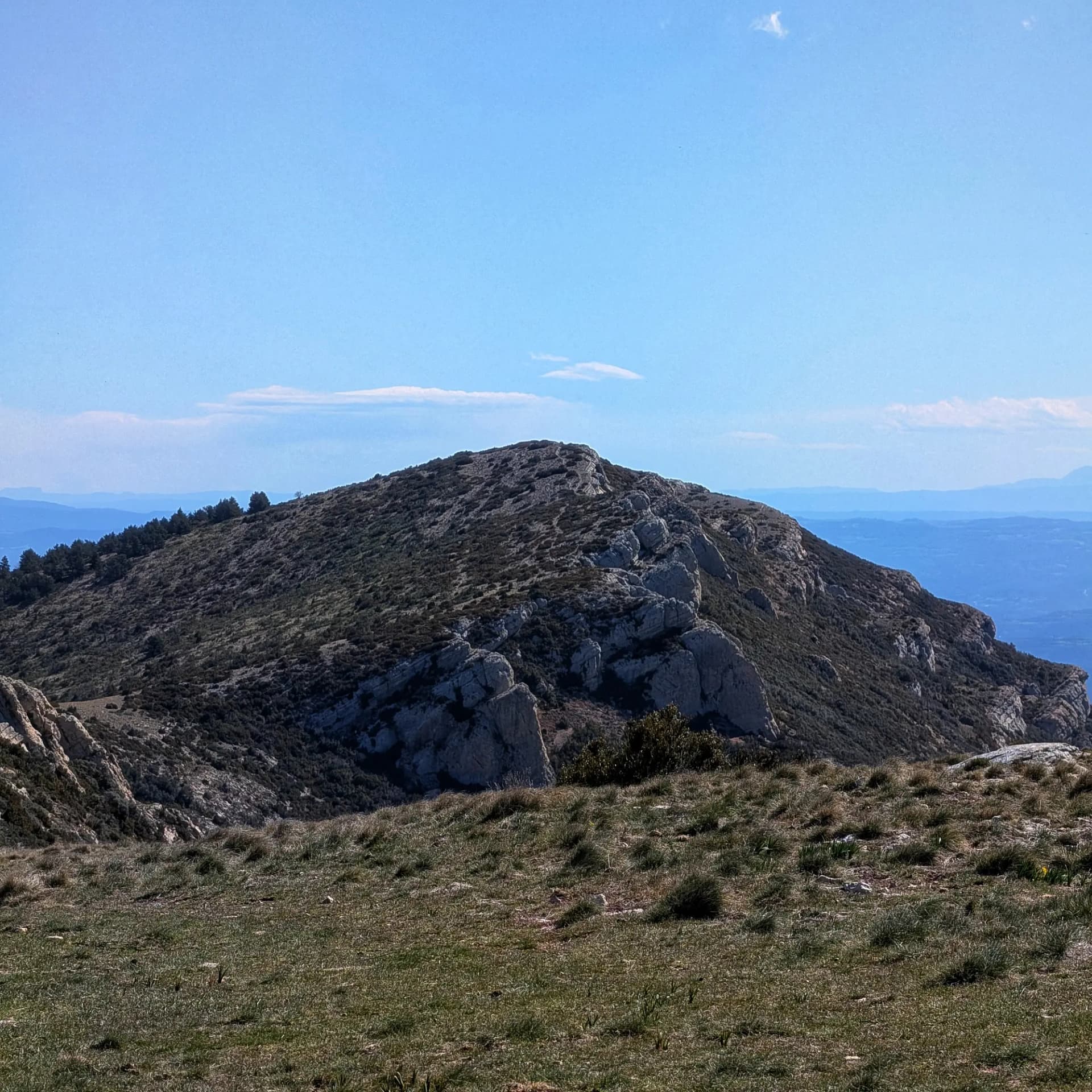 Tossal de Mirapallars rocoso y con matorral bajo cielo azul, con vistas lejanas a la Plana de Urgell.