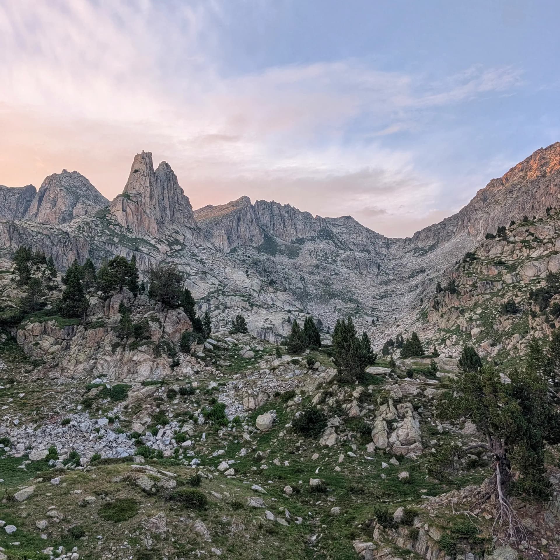 Vista panoràmica de pics muntanyosos escarpats i una vall rocosa amb arbres i herba sota un cel de capvespre.