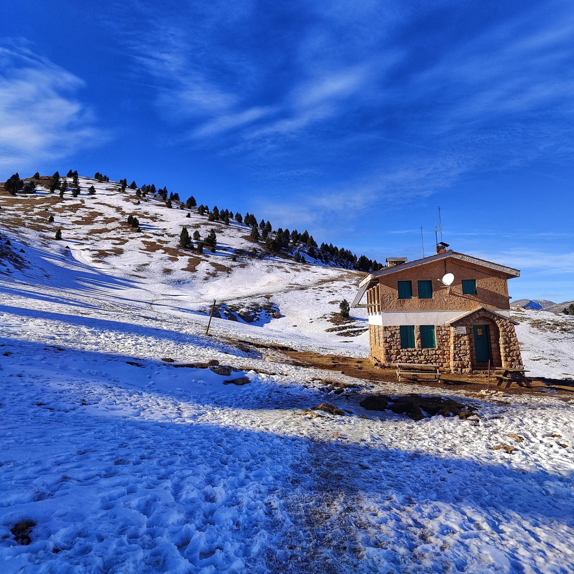 Refugio de montaña de piedra en la Serra d'Ensija, con nieve y pinos, bajo un cielo azul claro, en el ascenso al Cap de la Gallina Pelada.