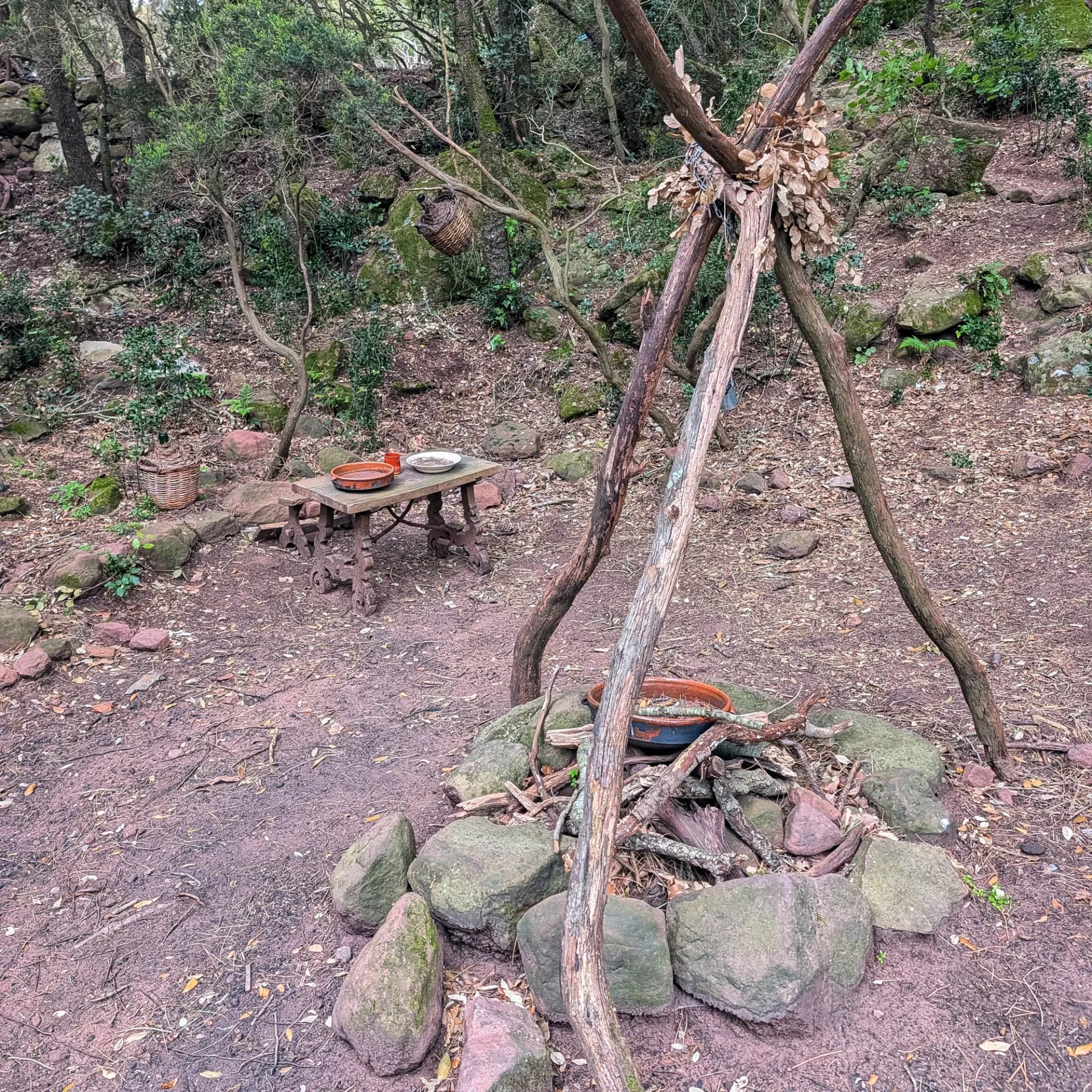 Clar del Bosc de les Bruixes amb foguera de pedra, taula rústica i trípode de branques. Entorn boscós i rocós.