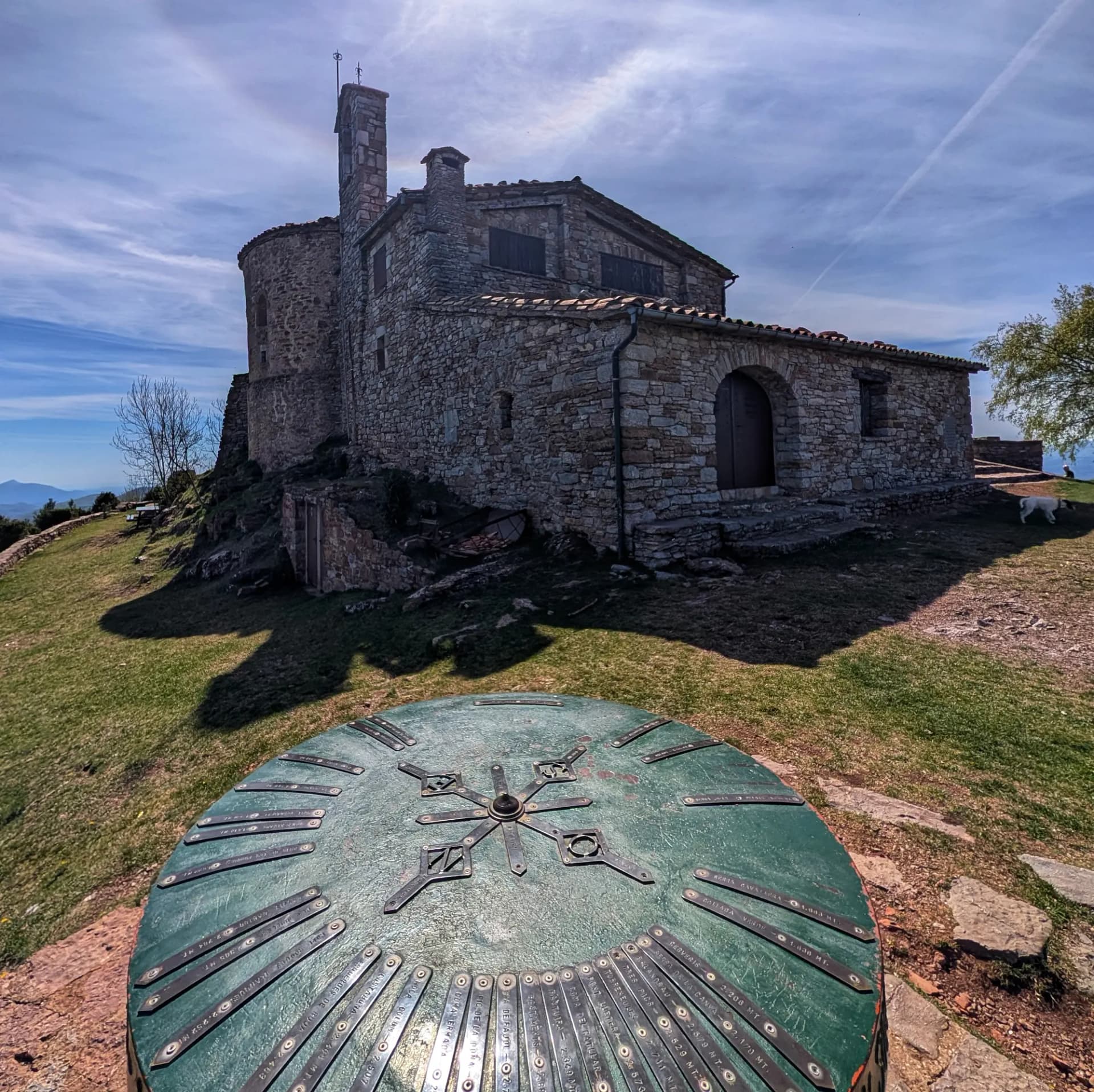 Ermita de Sant Antoni de pedra al cim, taula d'orientació verda en primer pla sota cel blau.