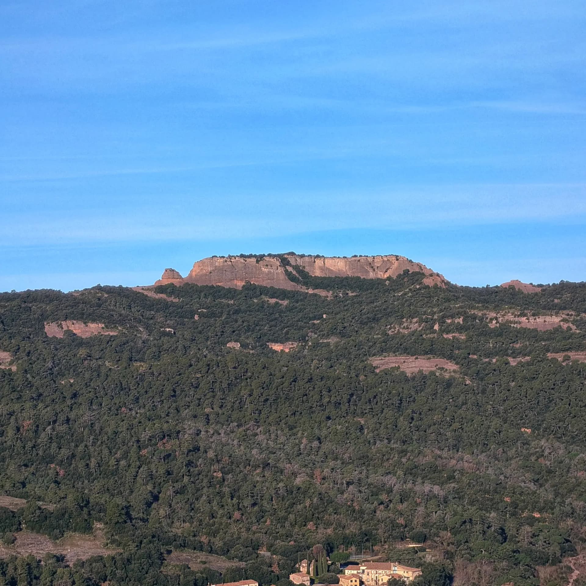 Vista panoràmica de la muntanya de Castellsapera amb densos boscos i un petit nucli de cases sota cel blau clar.