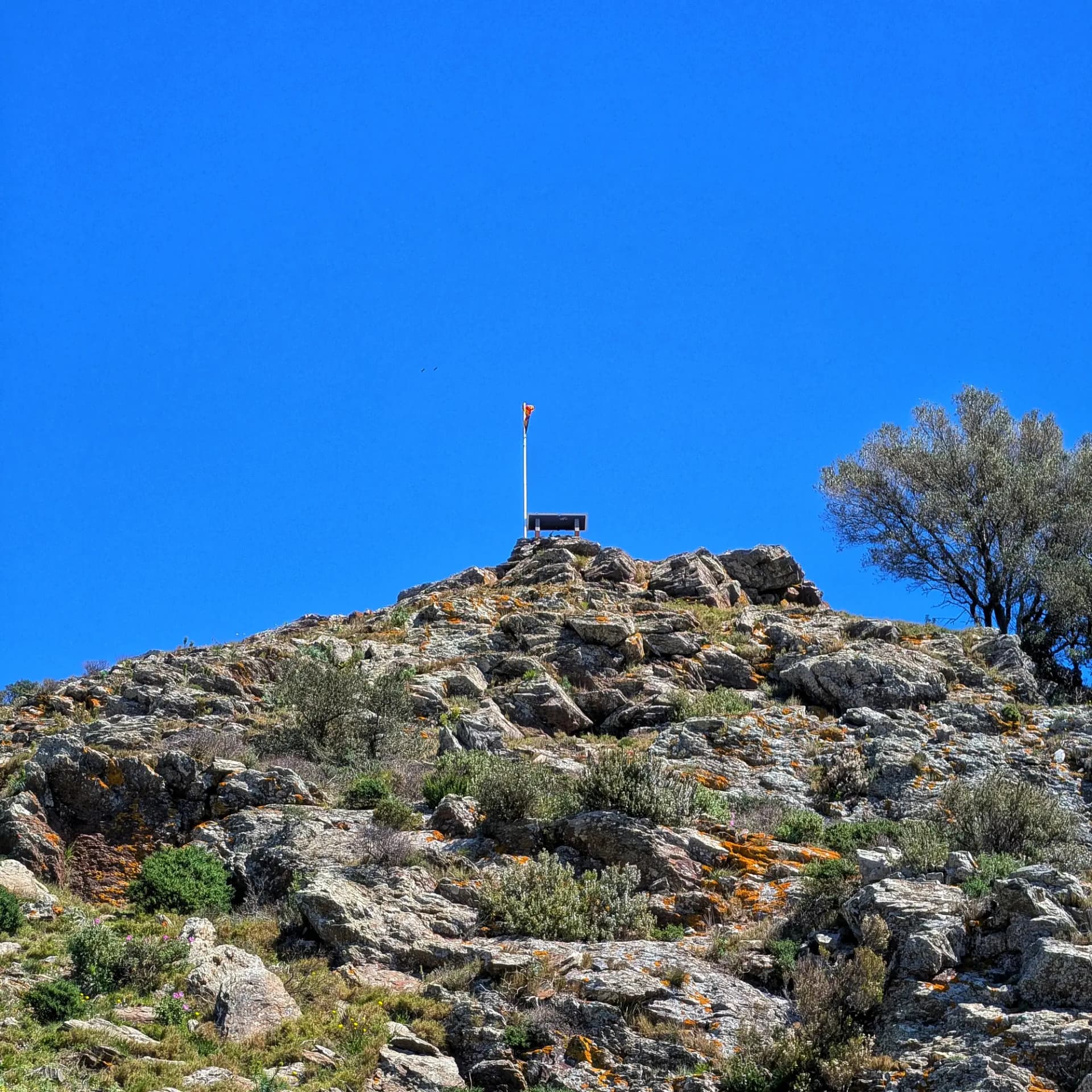 Cim rocós del Puig de l'Àliga amb bandera espanyola i plataforma d'observació sota un cel blau brillant.