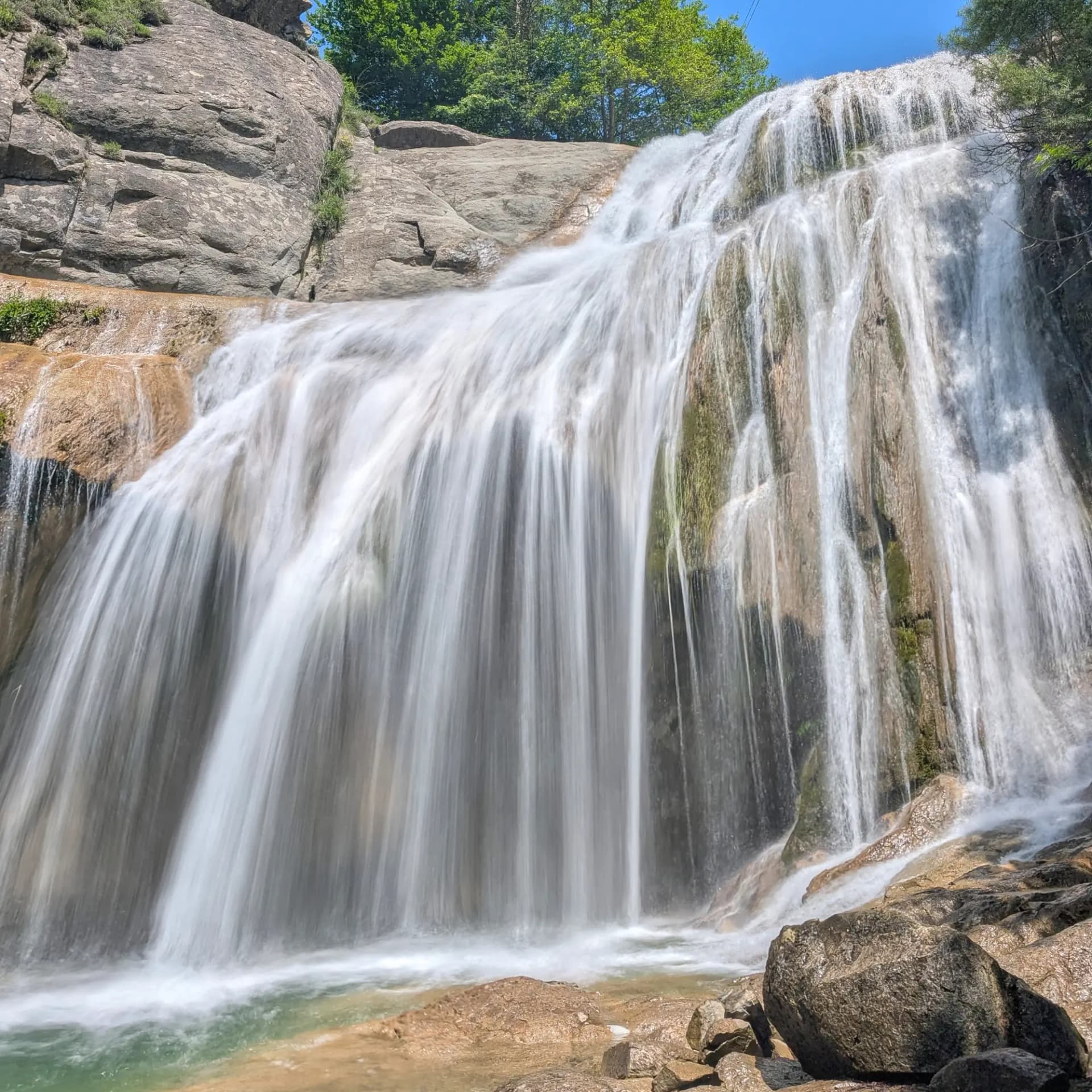 Large silky waterfall flowing down a rock face, surrounded by green foliage and a clear blue sky.