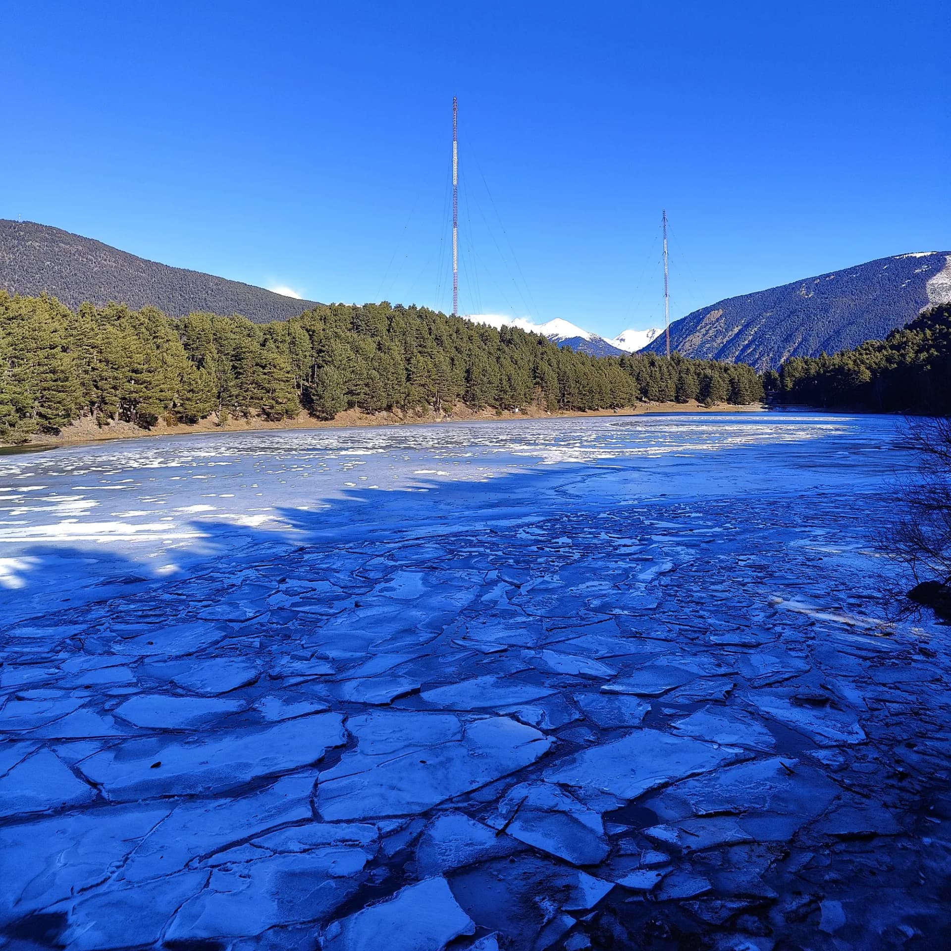 Frozen Estany d'Engolasters, with pine forests, snow-capped mountains, and communication towers under a clear blue sky.