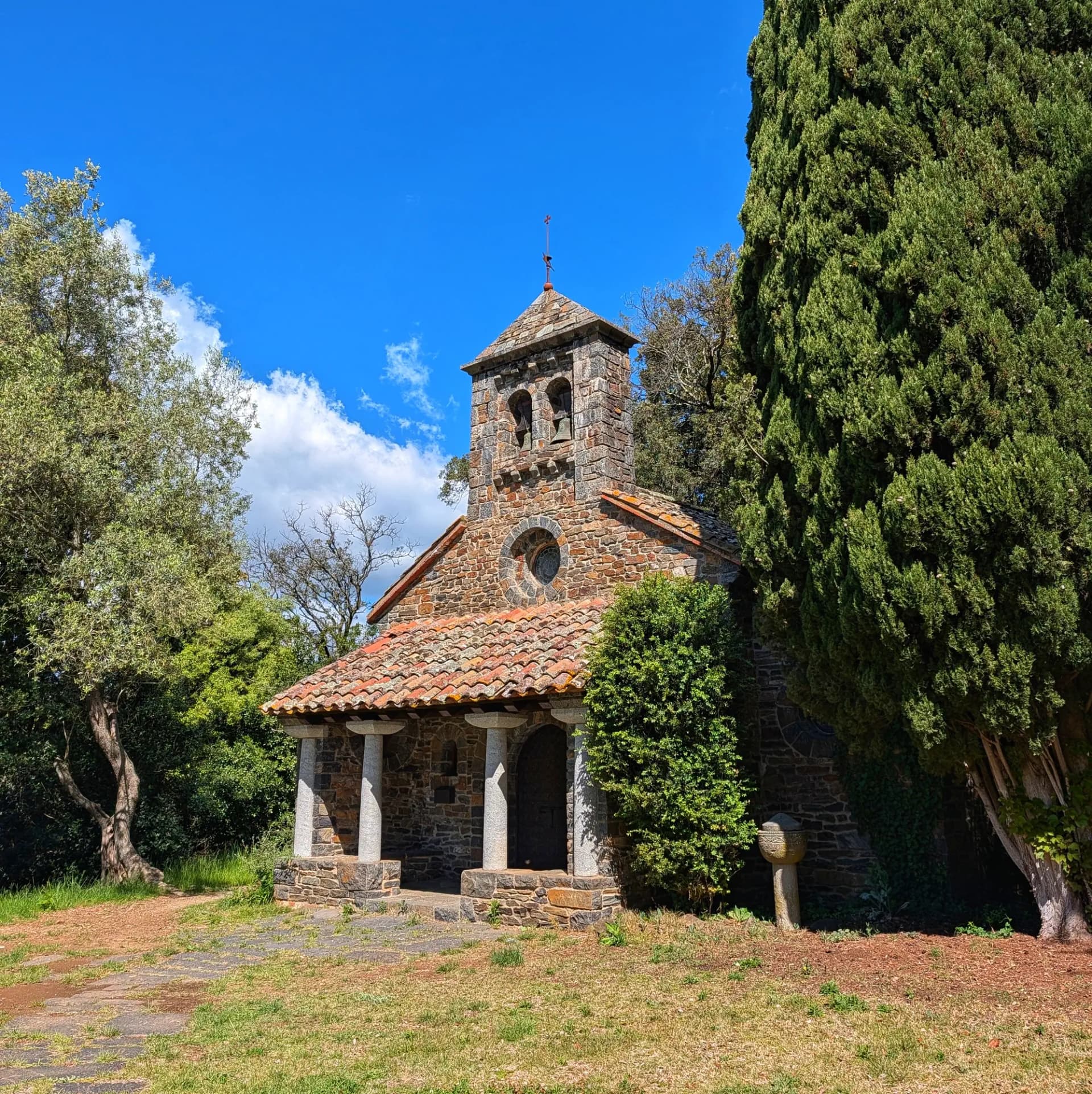 Ermita de pedra amb campanar, teulada de teula, porxo i òcul, envoltada d'arbres sota cel blau.