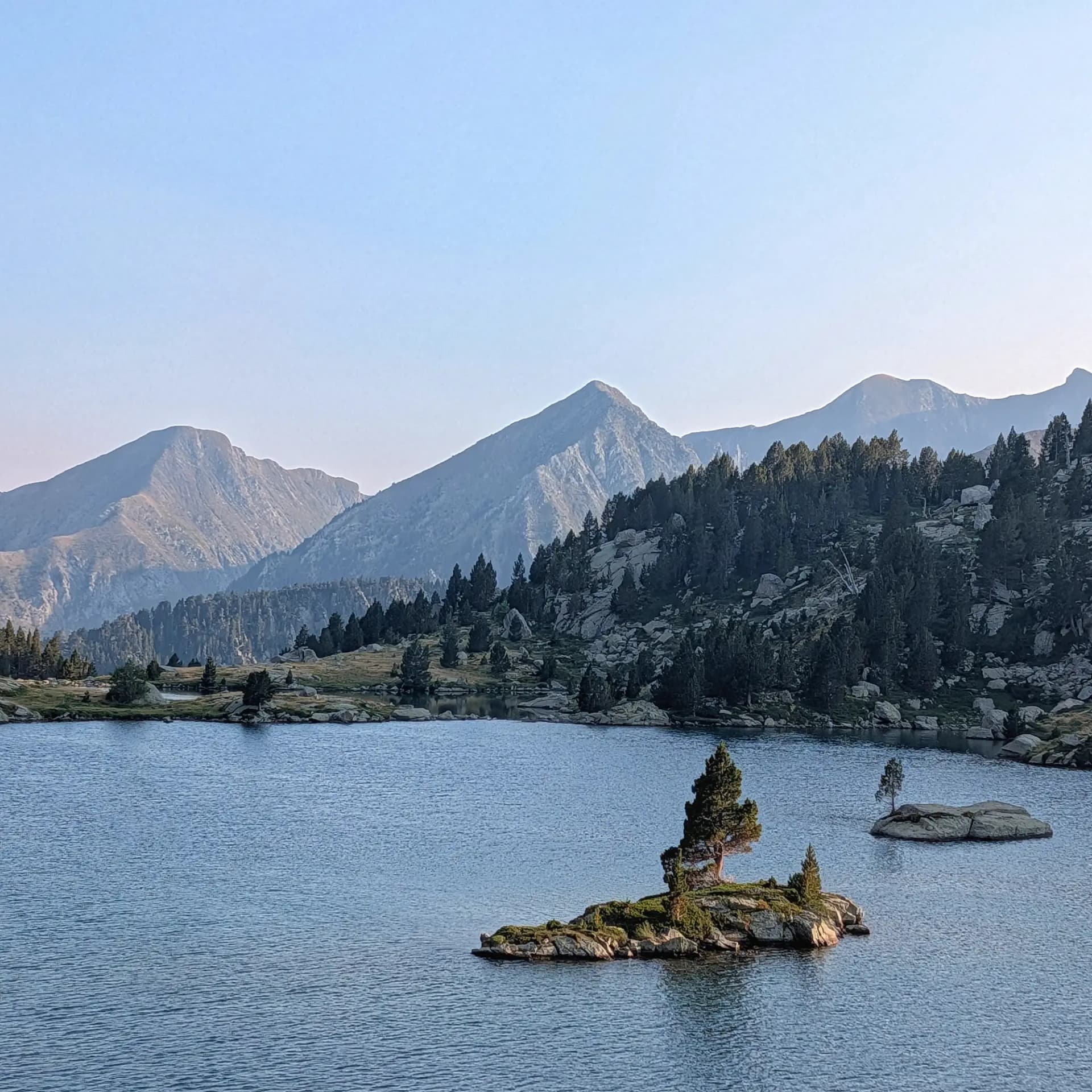 Llac de muntanya amb dues illes rocoses, pins, bosc dens i cims boirosos sota un cel blau clar.