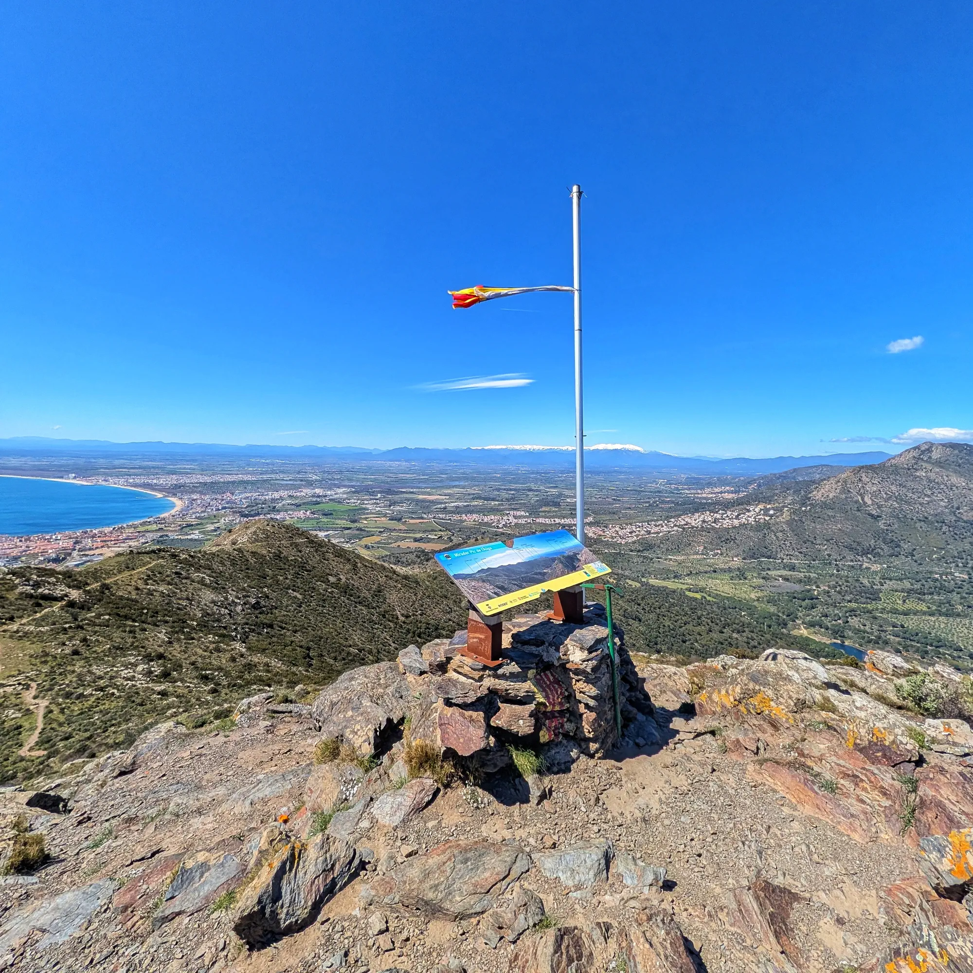 Vista panoràmica des del cim del Puig de l'Àliga amb plafó informatiu, costa, ciutat i muntanyes nevades.