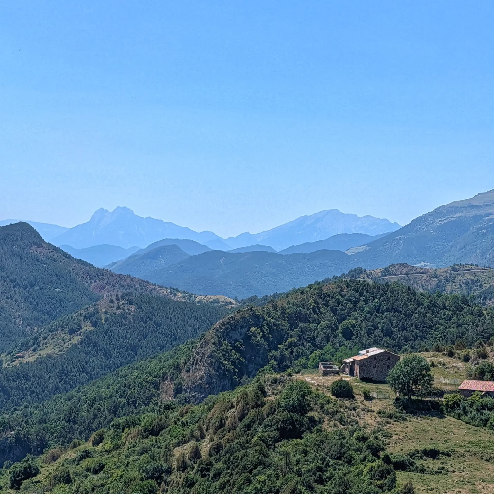 Panoramic view from Costa Pubilla, showcasing the lush green foothills leading to the iconic two peaks of Pedraforca (Pollegó Inferior 2445m, Pollegó Superior 2506m) and the Cadí range in the distance. Mountain landscape with layers of green hills, a stone house, and hazy blue distant peaks of Pedraforca and Cadí.