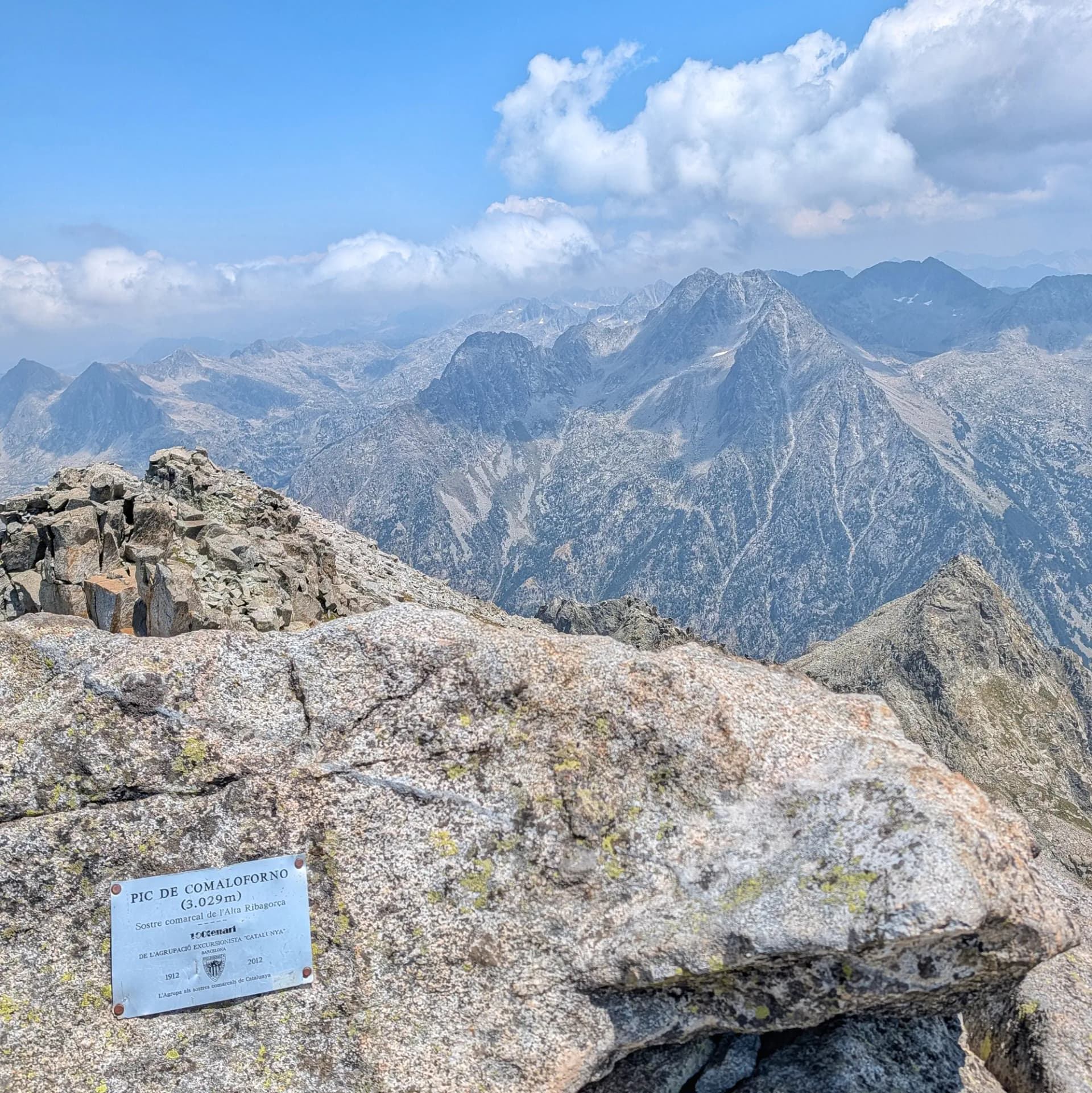 Placa metàl·lica en un cim rocós amb vista a una vasta serralada sota un cel blau parcialment ennuvolat.