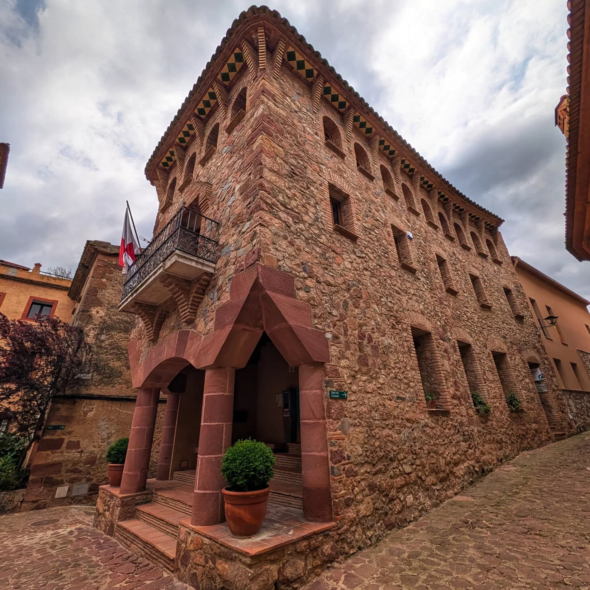 Edifici de l'Ajuntament de l'Argentera, amb façana de pedra i maó, balcó i teulada decorada, sota un cel ennuvolat.