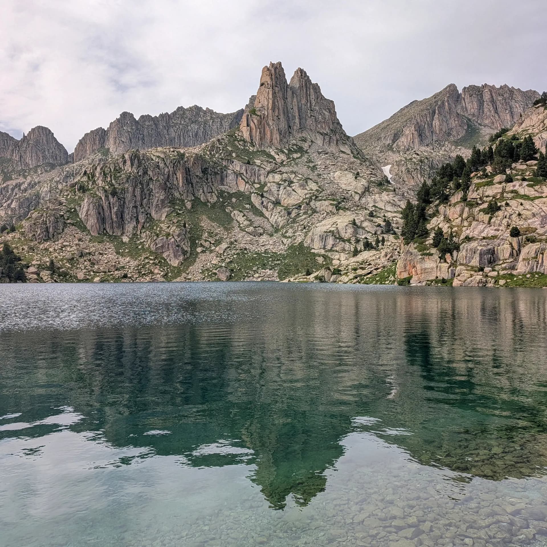 Llac de muntanya cristal·lí que reflecteix pics de granit i vessants rocosos sota un cel ennuvolat. Roques subaquàtiques visibles.