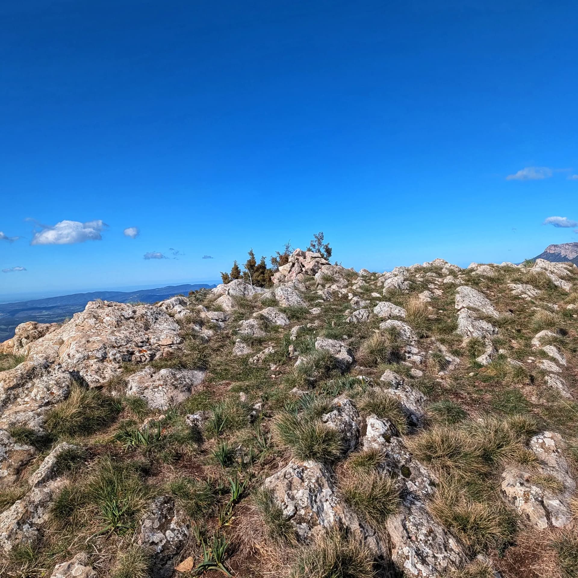 Rocky summit with sparse vegetation, a cairn, and clear blue sky with scattered clouds.