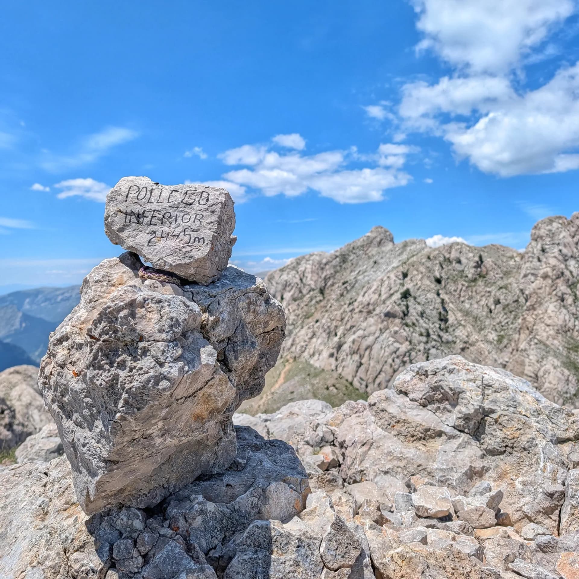 Summit cairn marked 'POLLEGO INFERIOR 2445m' on a rocky peak, with rugged mountains and blue sky with clouds.