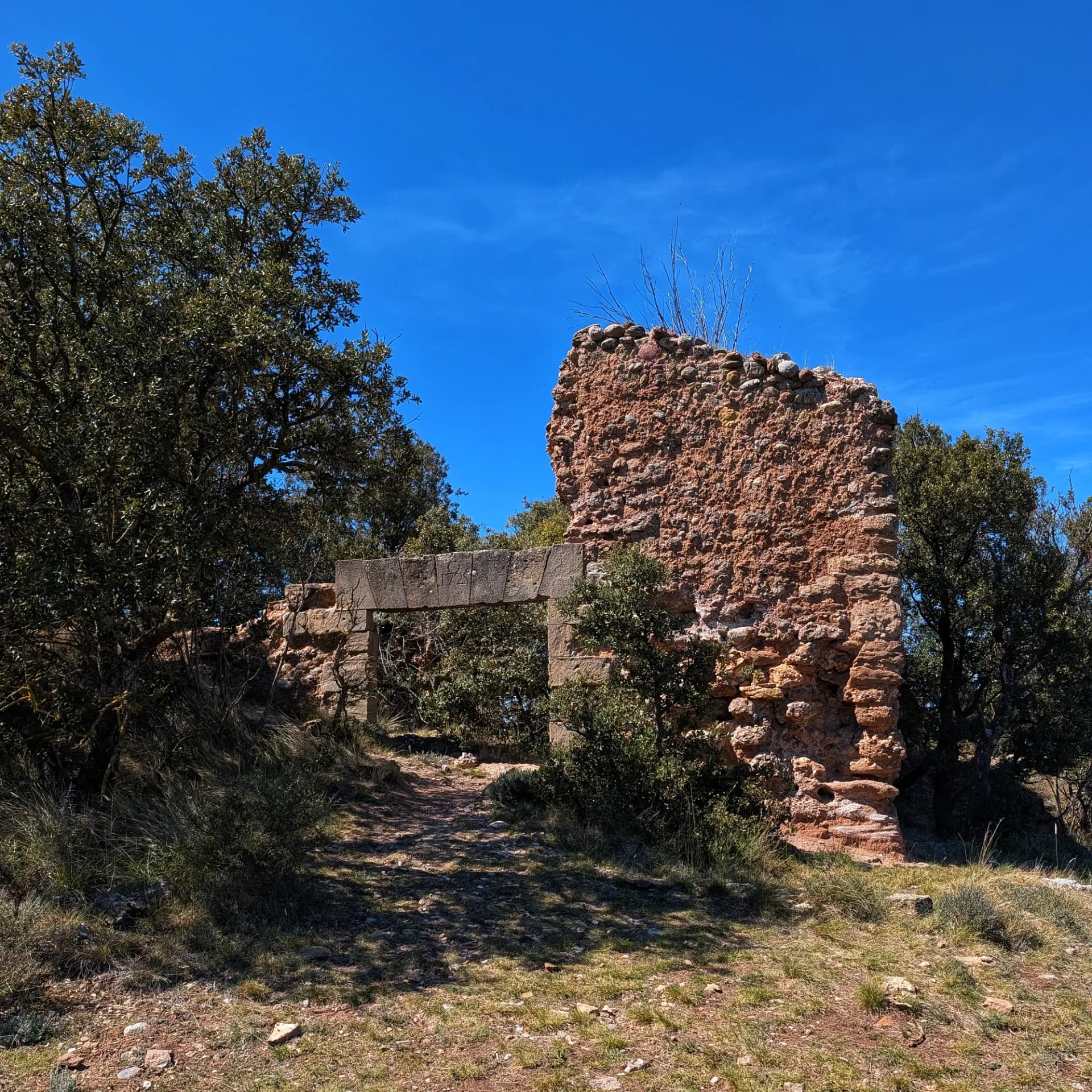Ruïnes d'una paret de pedra vermellosa d'ermita amb un arc, envoltades d'arbres verds i herba sota un cel blau clar.