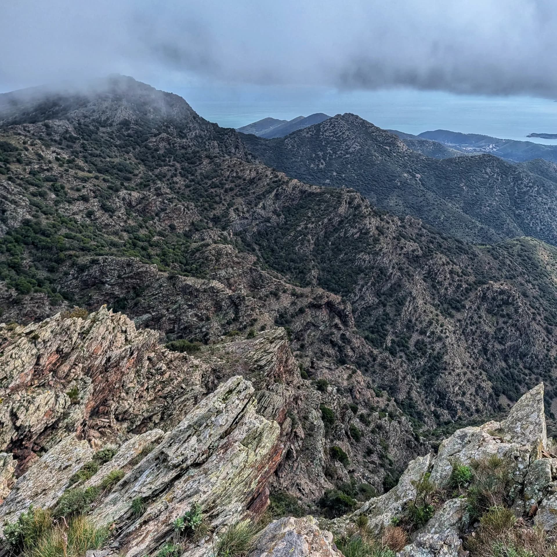 Vistes de muntanyes rocoses i vegetació esparsa sota un cel ennuvolat, amb el mar a l'horitzó.