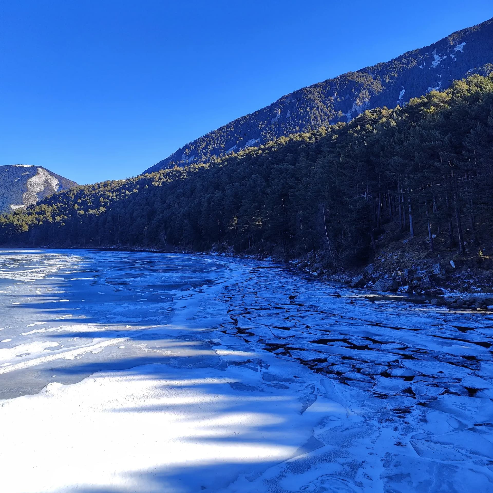 Partially frozen mountain lake with cracked ice, dense pine forest, and snow-dusted peaks under a clear blue sky.